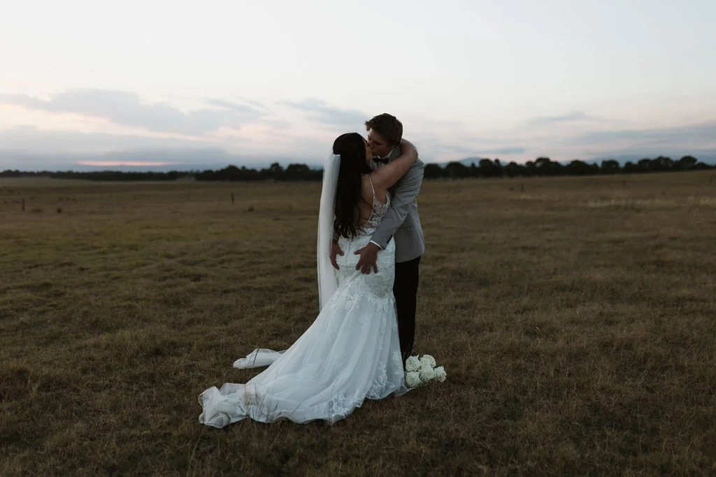 A newlywed couple embracing in a grassy field during sunset, with the bride in a lace wedding dress and the groom in a suit.