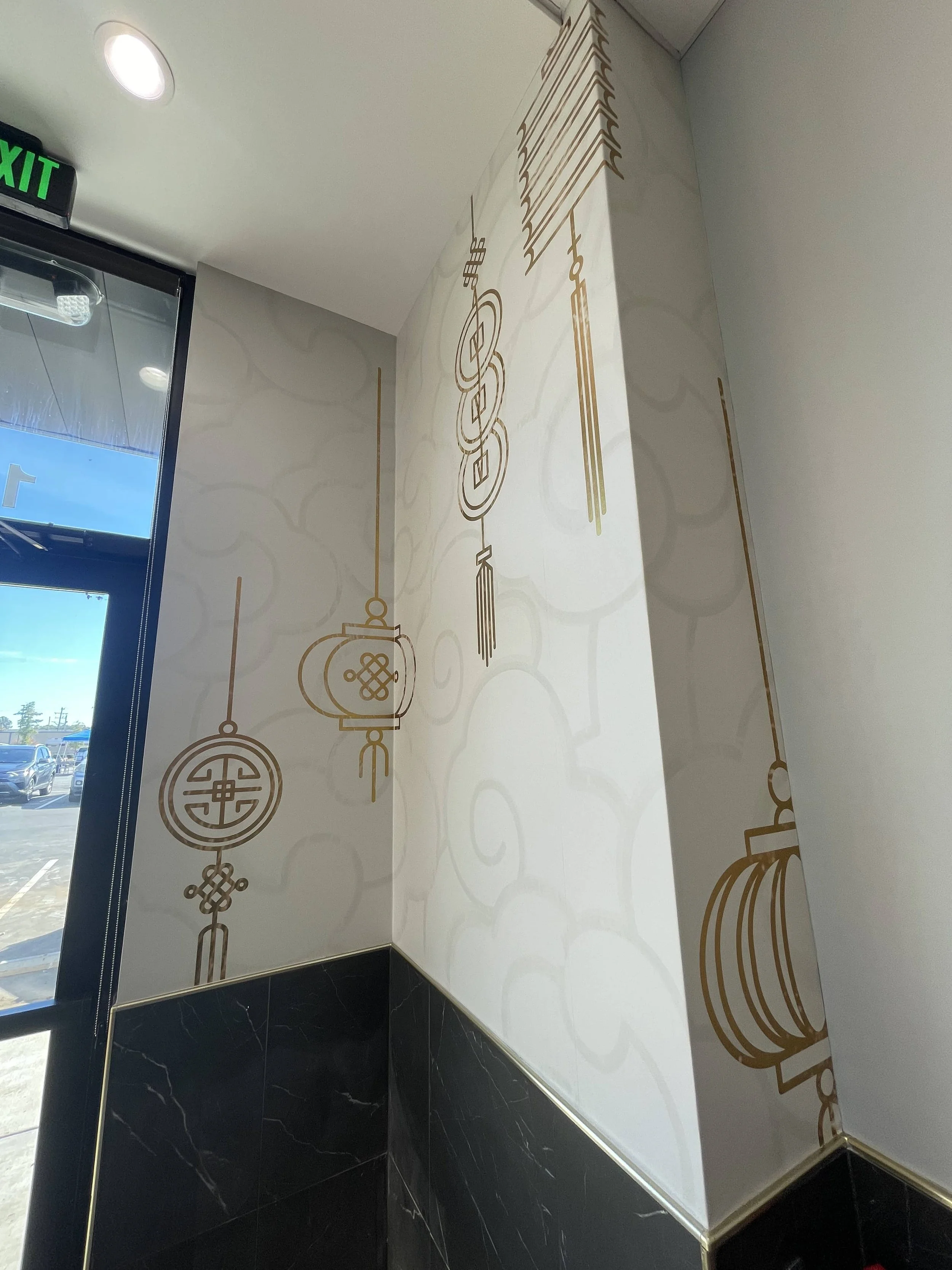 Interior corner of a room with decorative wall art featuring traditional Chinese symbols and lantern designs, partially illuminated by natural light from a nearby glass door.