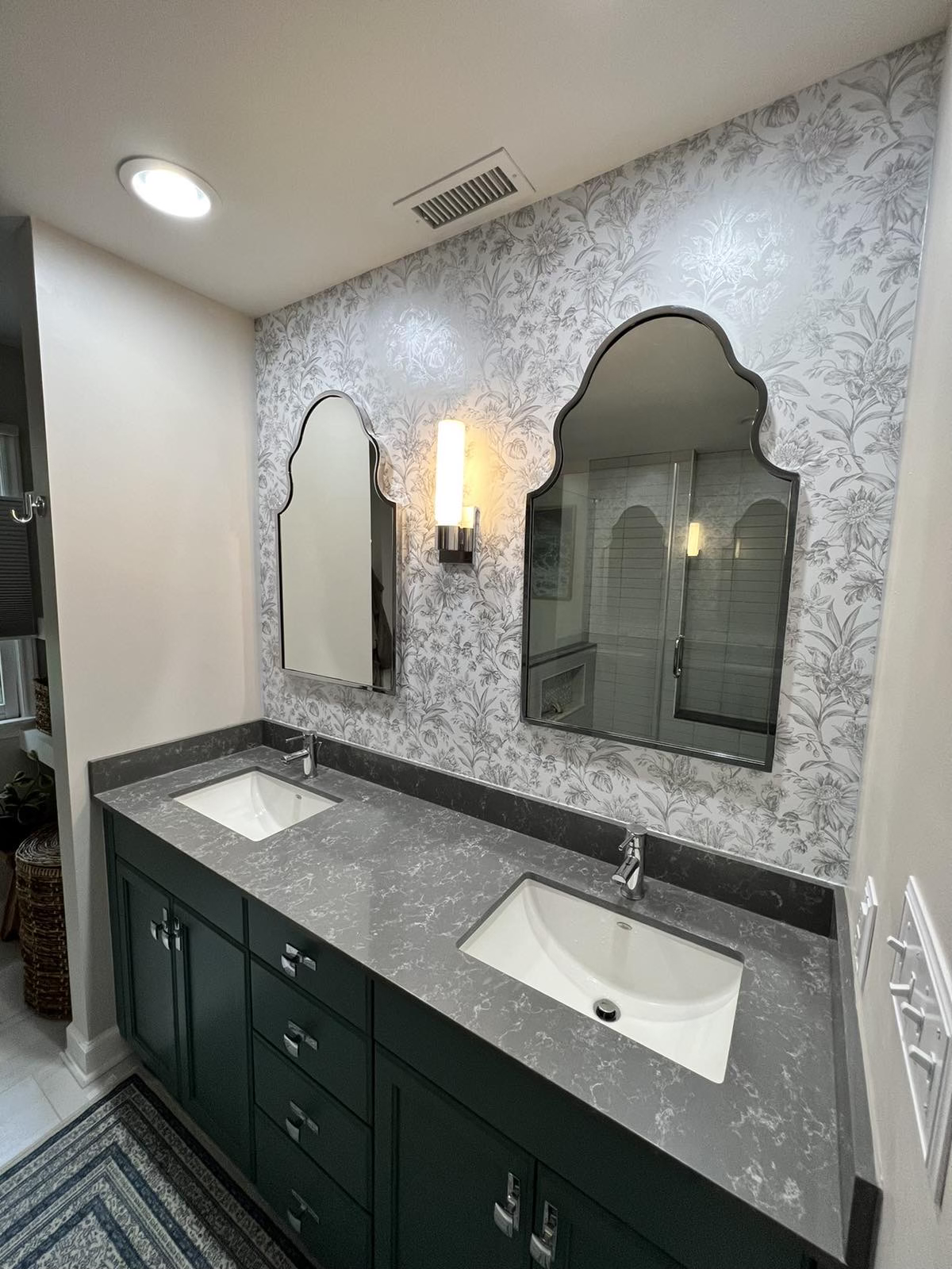 Double bathroom vanity with dark gray marble countertop, two rectangular sinks, and two arched mirrors on floral wallpapered wall, with wall-mounted light fixture. Wallpaper instalating in Charlotte and near