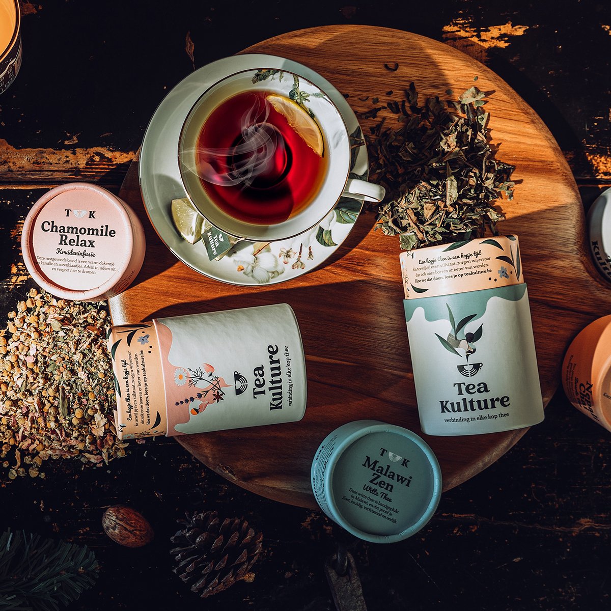 A cup of steaming tea with lemon slices on a saucer, surrounded by tea packages labeled "Tea Kulture," "Chamomile Relax," and "MalawiZen," on a wooden surface with dried herbs, pine cones, and tea leaves.