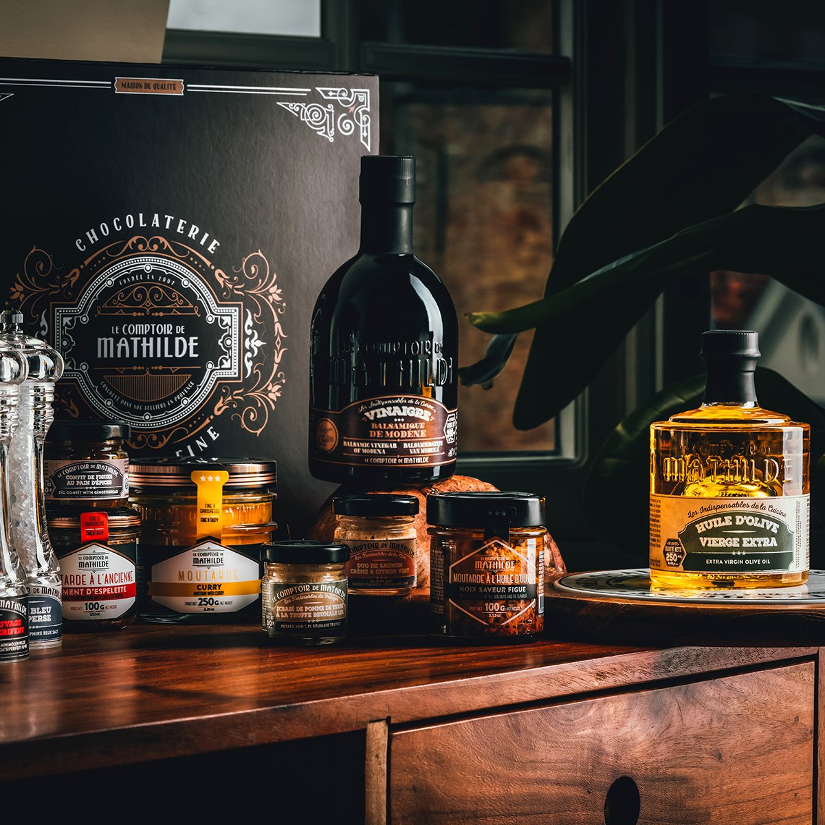Selection of gourmet food products including jars, bottles, and containers of mustard, vinegar, and olive oil on a wooden table, with decorative packaging and a large black box in the background.