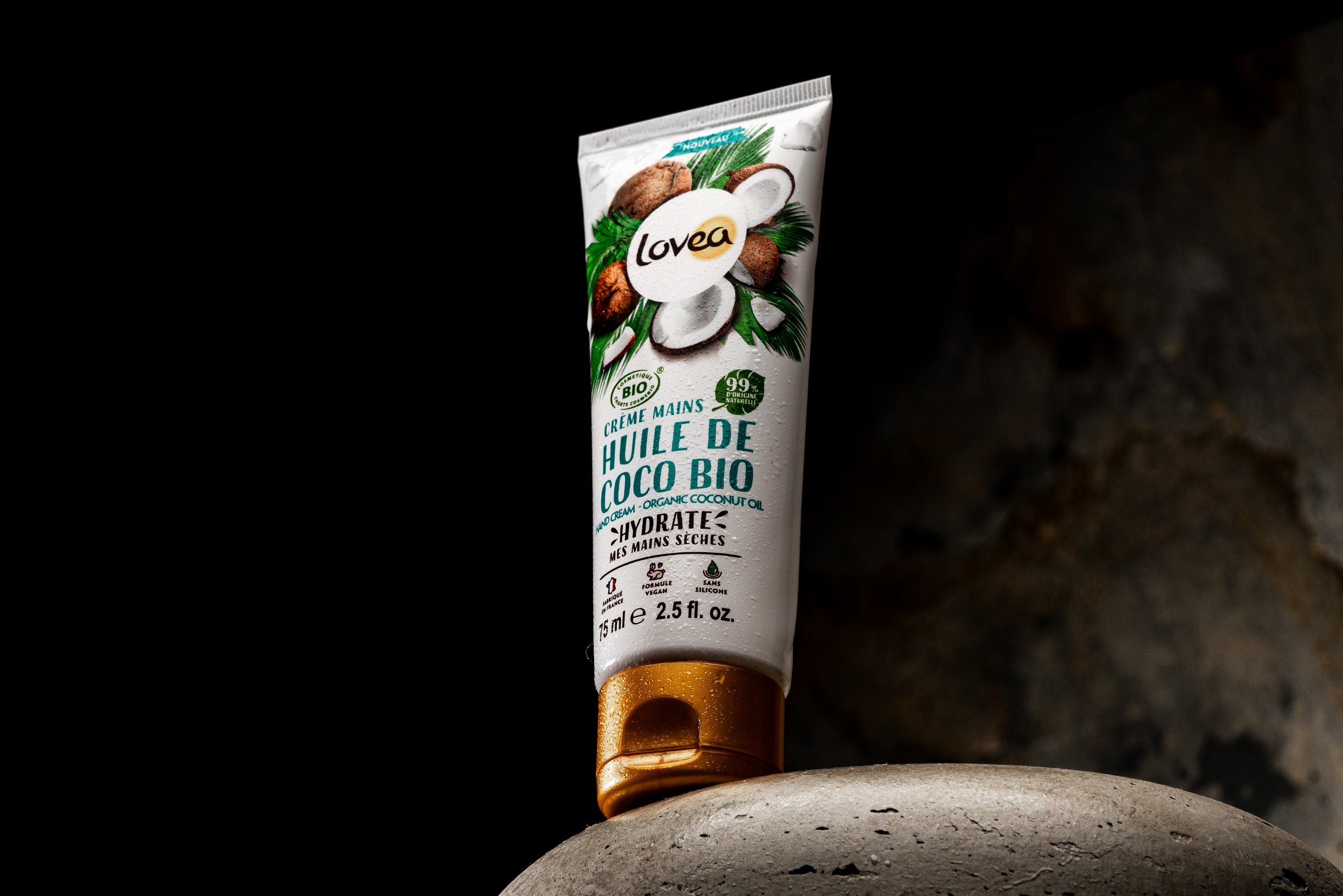 A tube of Lovea organic coconut oil hand cream standing upright on a curved stone surface, with a dark background.