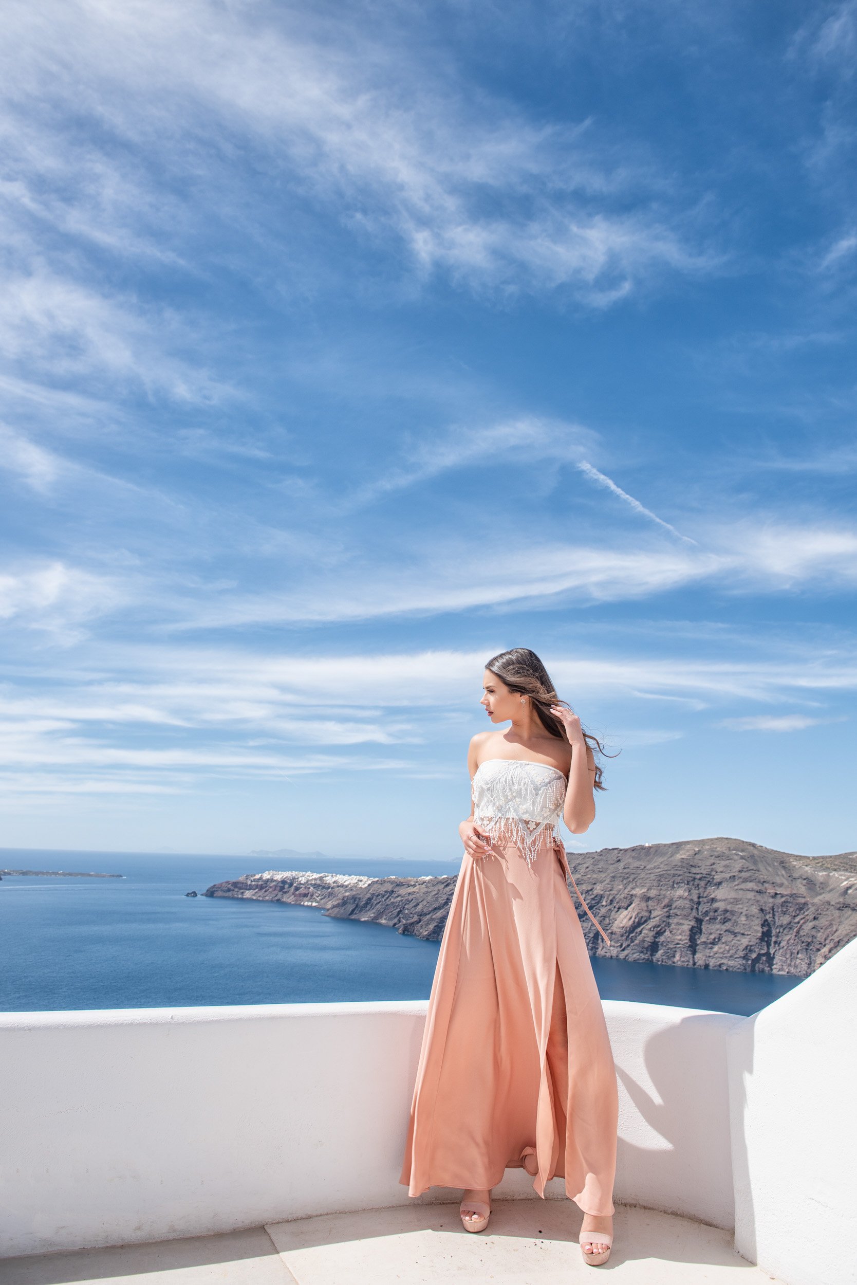 A woman in a strapless white top and long peach skirt stands on a white terrace with a view of the ocean and cliffs under a bright blue sky with wispy clouds.
