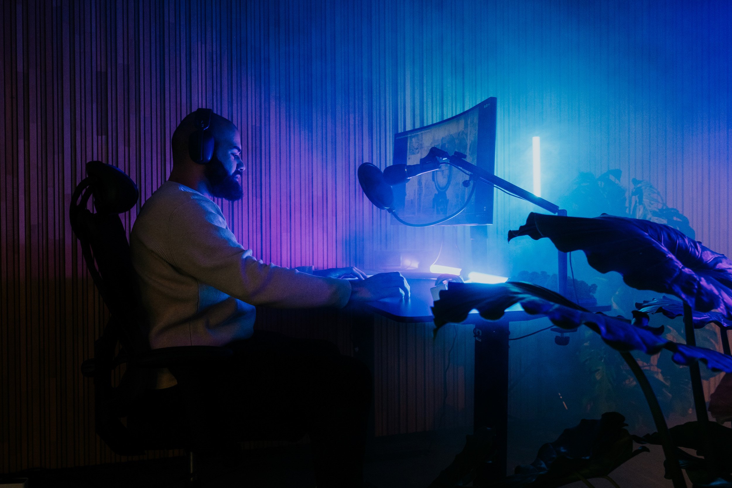 A man sitting in a gaming chair, wearing headphones, playing video games at a desk with multiple monitors, illuminated by blue and purple LED lighting, with plants nearby.