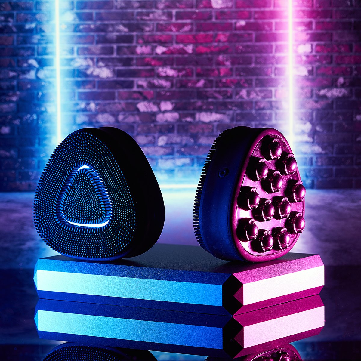Two massage rollers, one blue triangular textured and one purple with spherical rollers, on a reflective surface against a brick wall illuminated with pink and blue neon lights.
