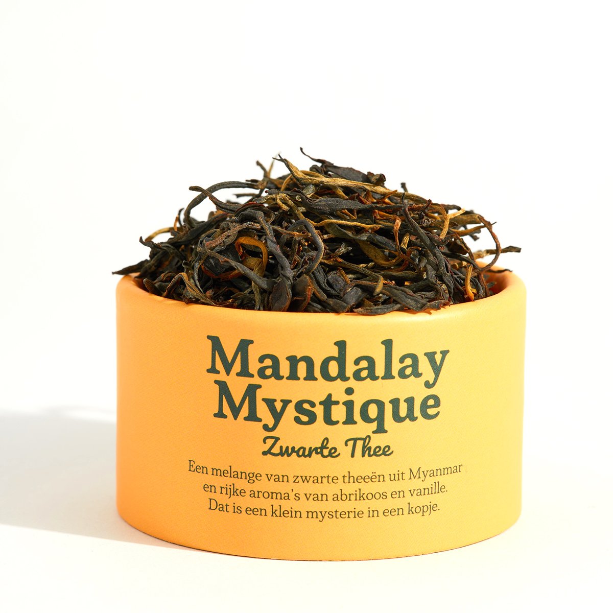 A yellow container filled with dried black tea leaves labeled "Mandalay Mystique Zwarte Thee," with Dutch text describing the tea's origins and flavors.