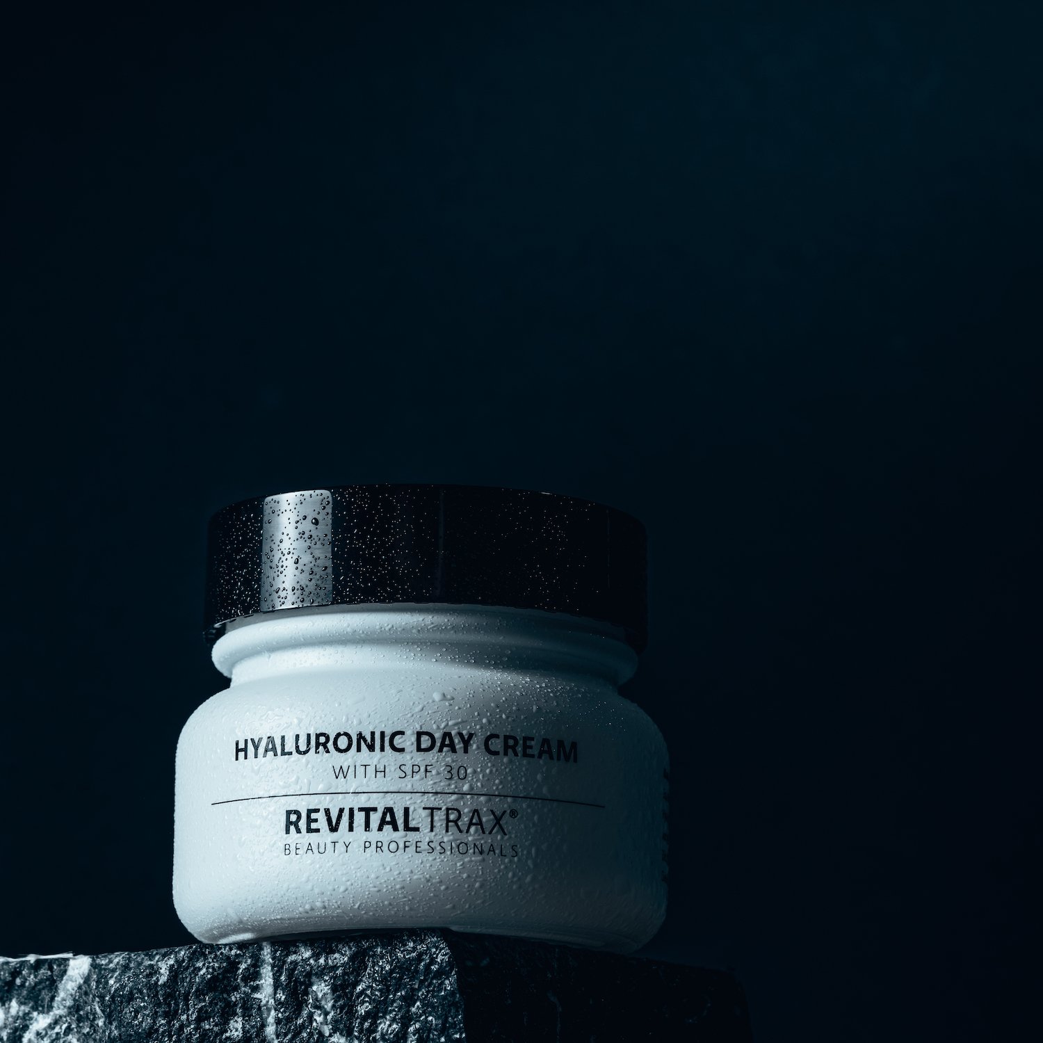 A white jar of Hyaluronic Day Cream with SPF 30 from RevitalTrax, showing water droplets on the surface, placed on a dark textured surface against a dark background.