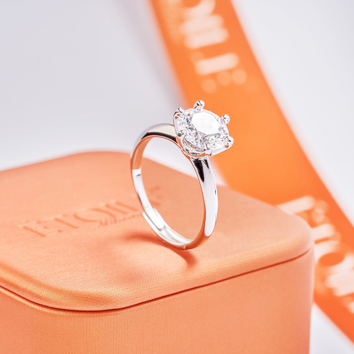 A silver ring with a large, clear gemstone, possibly a diamond, set in a prong setting. The ring is placed on an orange box with the brand name Hermès, and a blurry orange box or packaging is in the background.