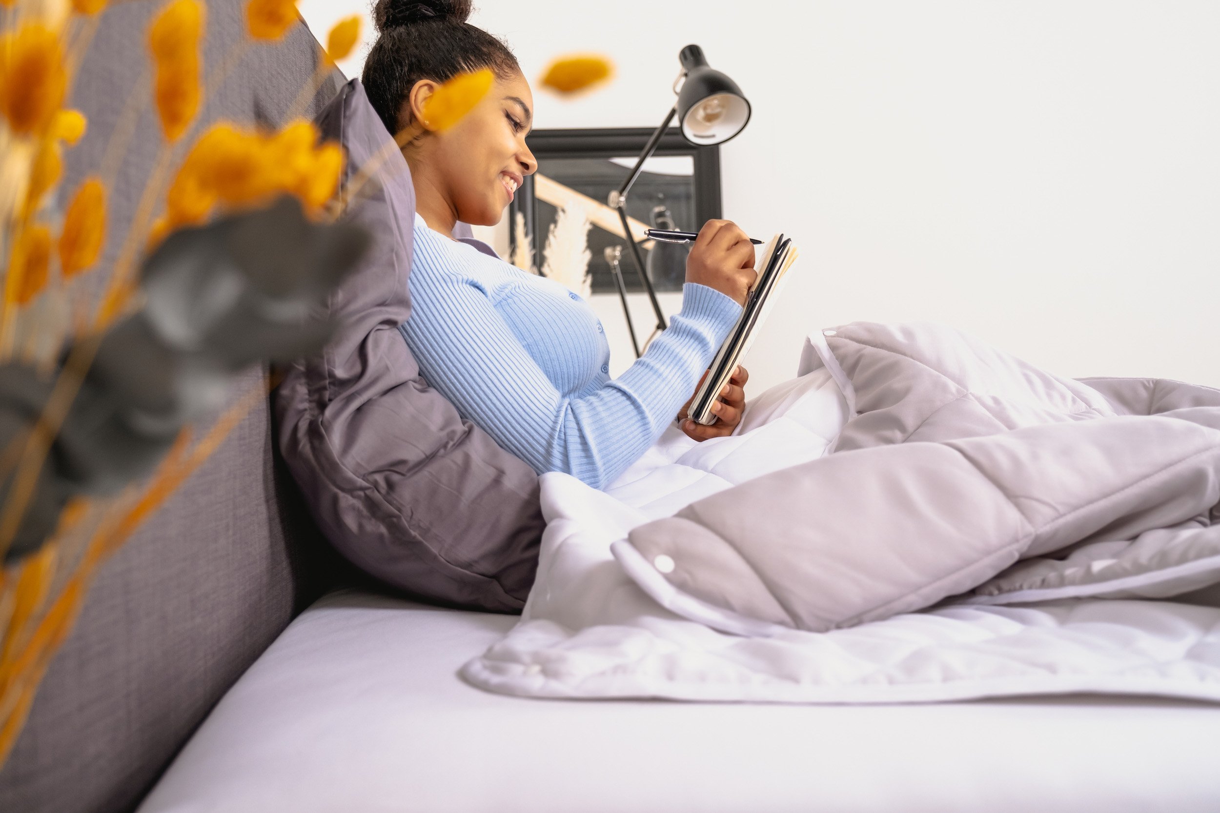 A woman with dark hair in a bun is sitting up in bed, looking at a notebook and smiling. She is covered with a gray comforter and there is a black framed mirror and a black desk lamp behind her. In the foreground, there are yellow and gray flowers ou