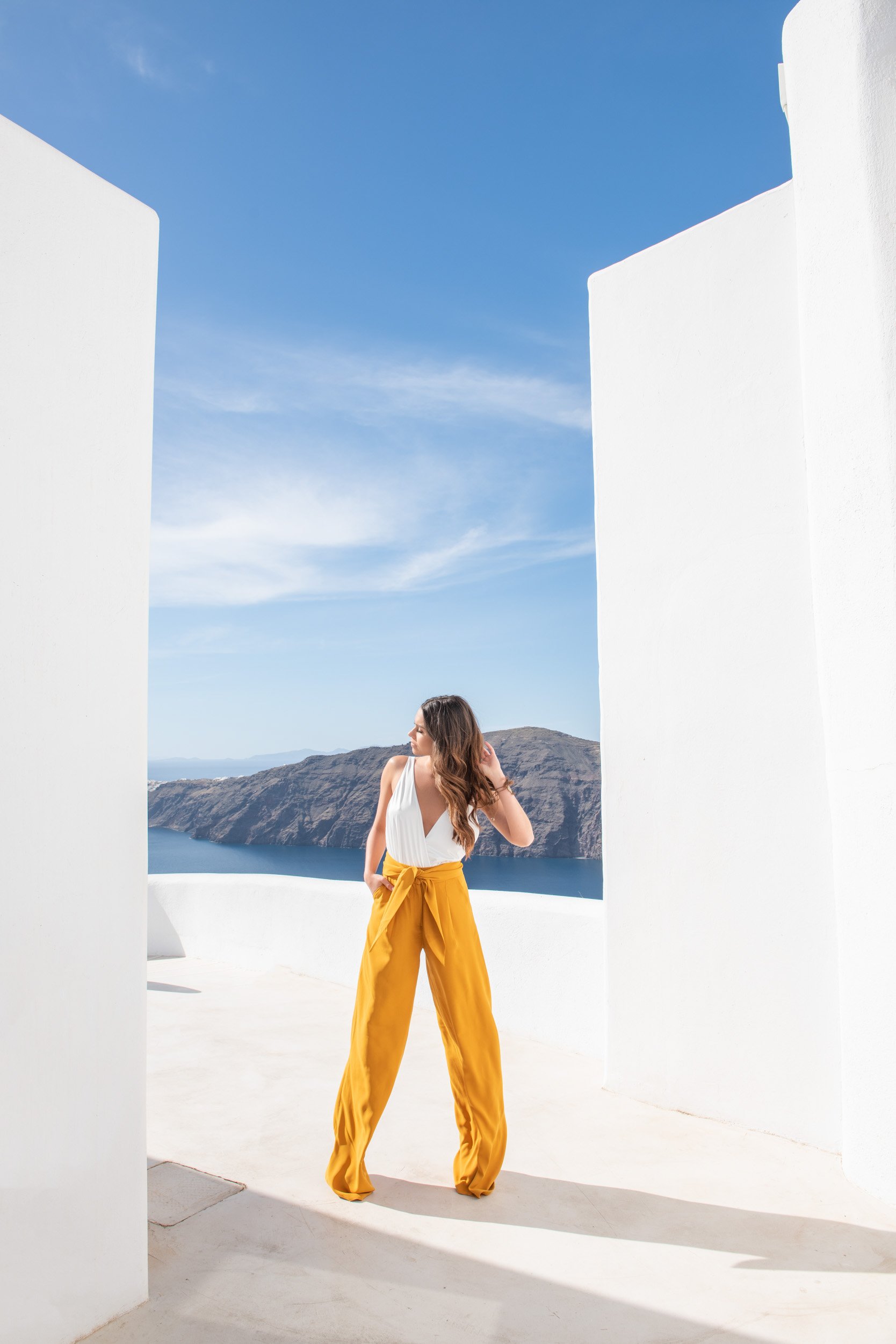 A woman with long brown hair stands outdoors between two white walls, overlooking a body of water and mountains under a blue sky with wispy clouds.