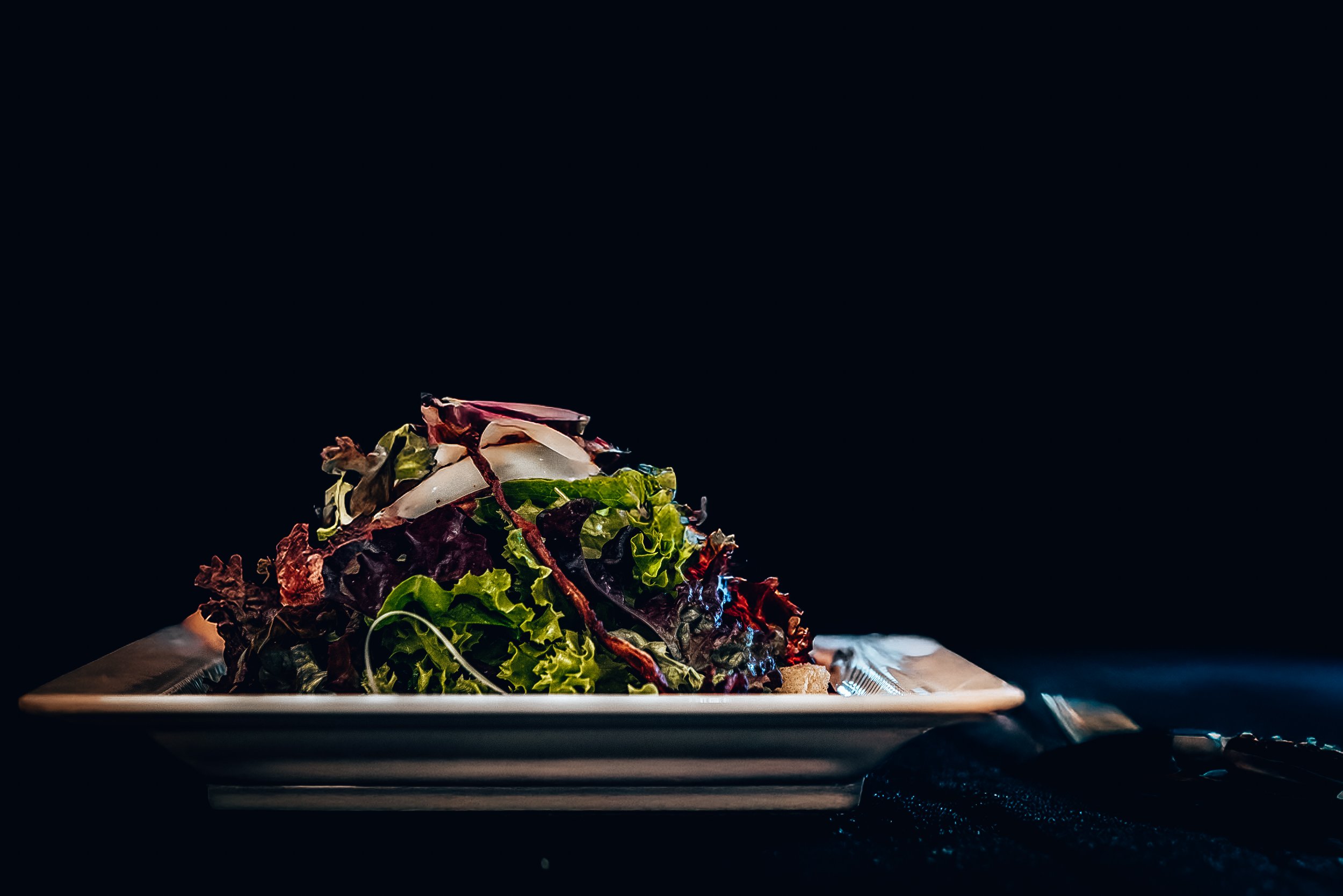 A plate of mixed green and purple leafy salad with shavings of cheese on top, served on a dark background with a silver fork and knife beside it.