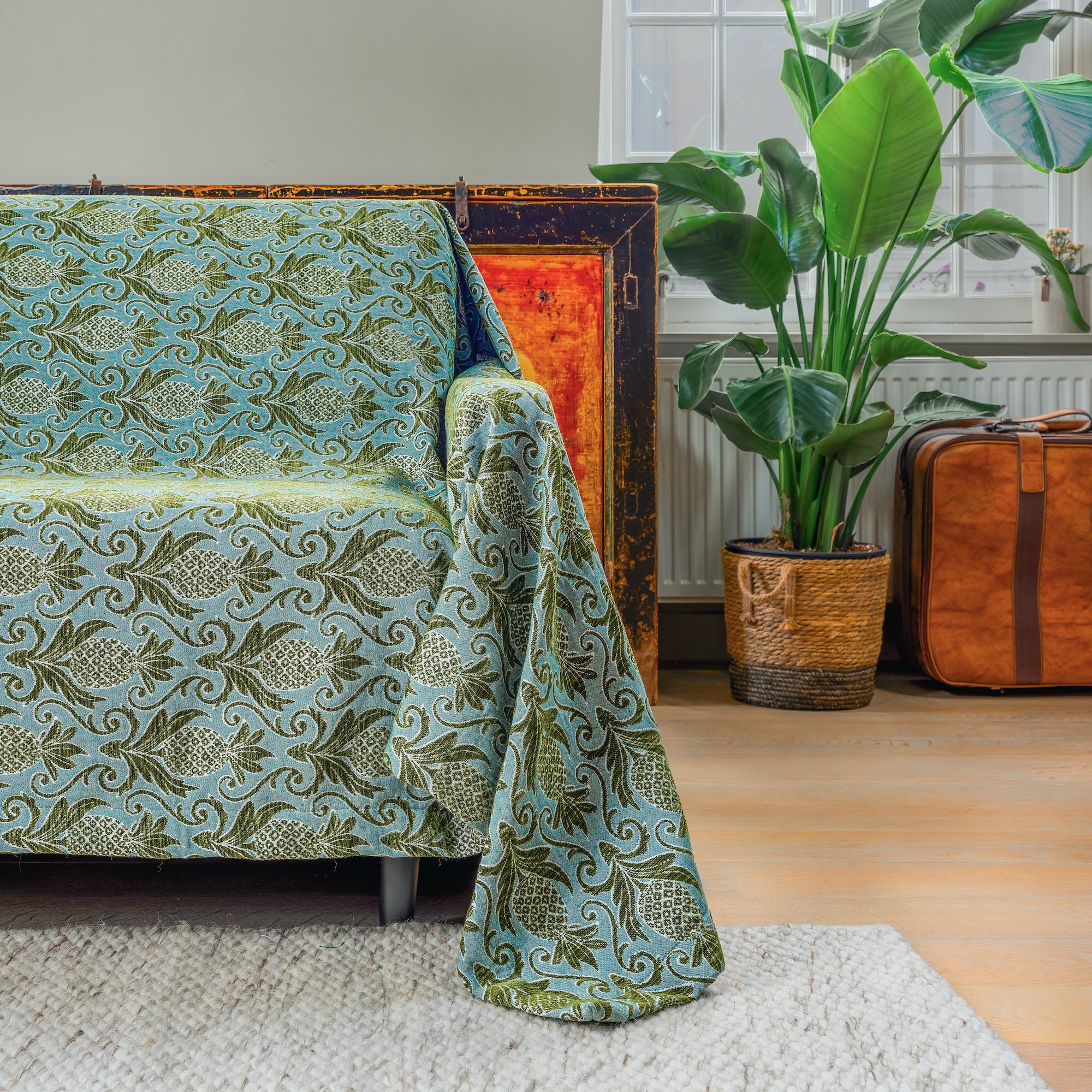 A living room corner with a patterned sofa cover, a large green potted plant, and brown suitcase near a window.