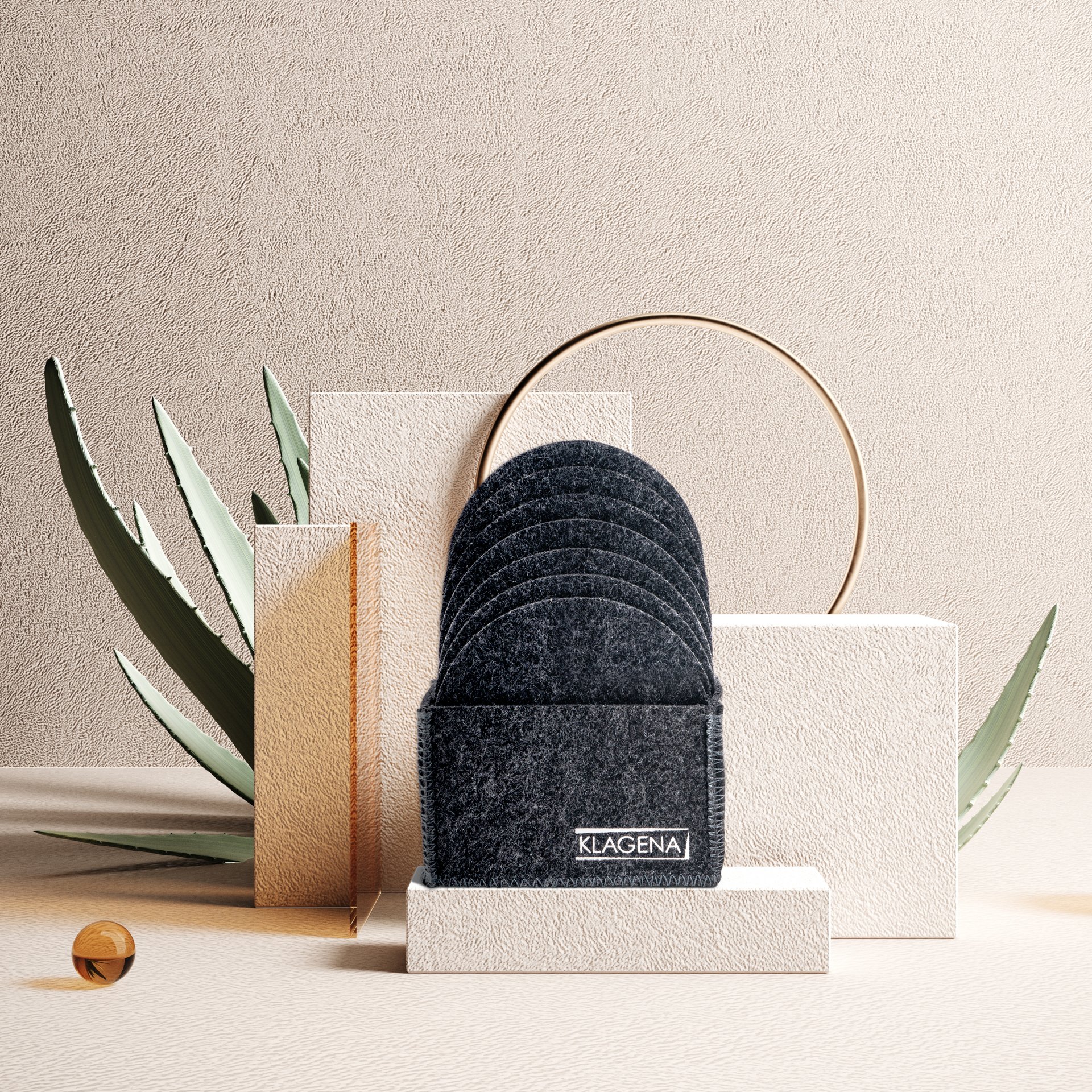 Stack of six black Klagen cleaning scrubbers on a white display stand, with abstract decorative elements including a gold circle, textured gold and white blocks, green leaves, and a small wooden sphere on a beige textured background.