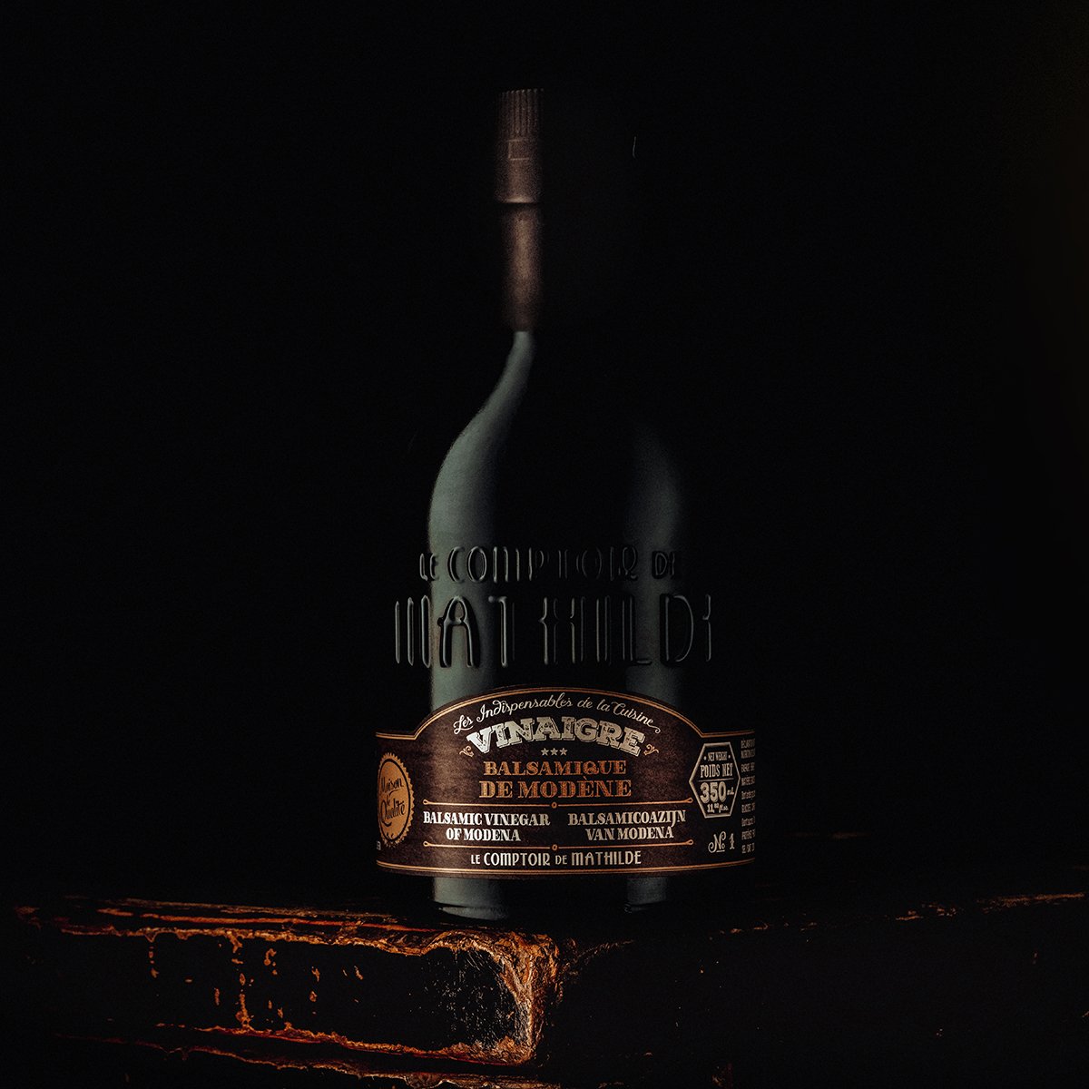A dark image of a bottle of balsamic vinegar from Le Comptoir de Mathilde on a rustic wooden surface.