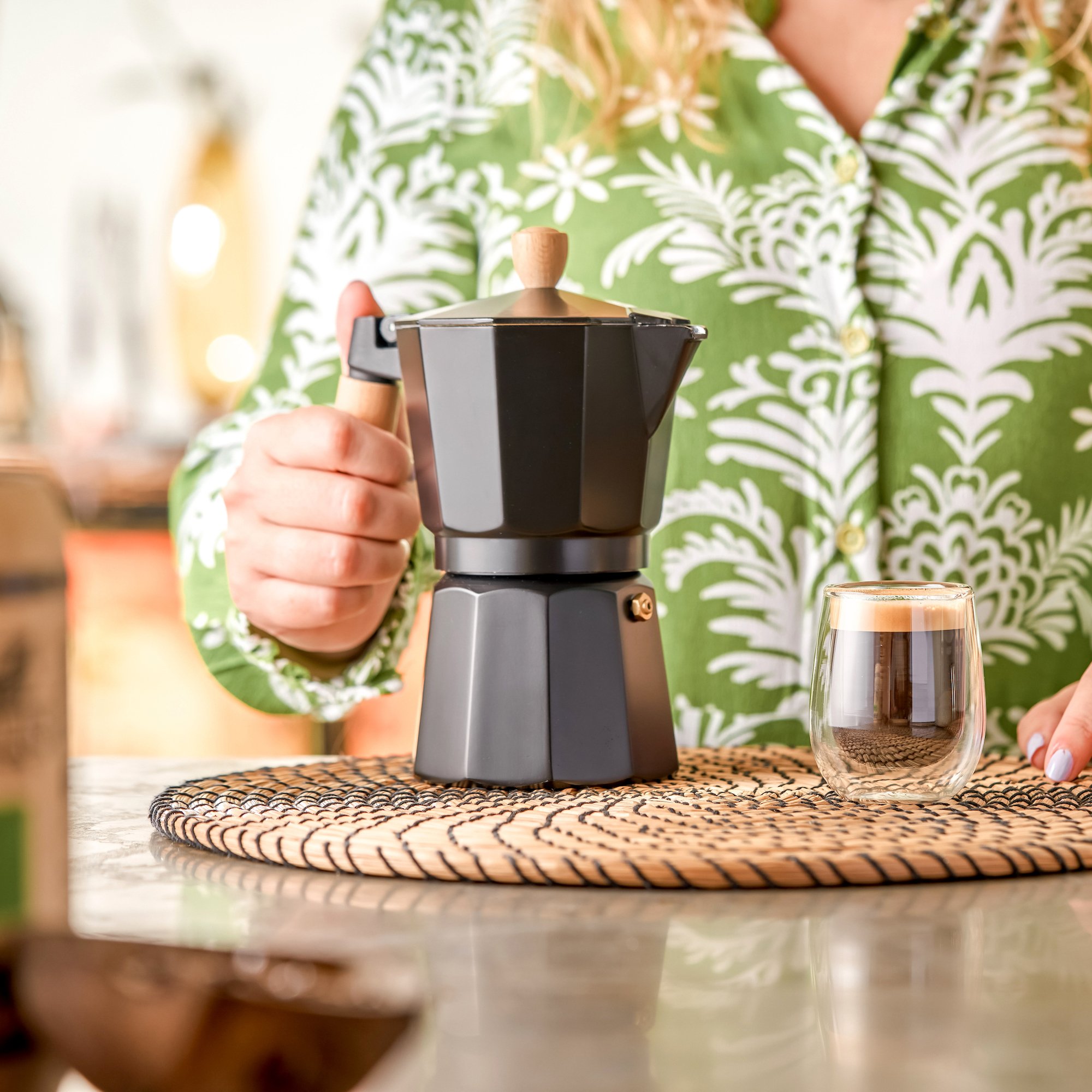 A person in a green and white floral shirt is preparing coffee with a moka pot on a woven placemat. There is a glass of water to the right of the moka pot on the table.