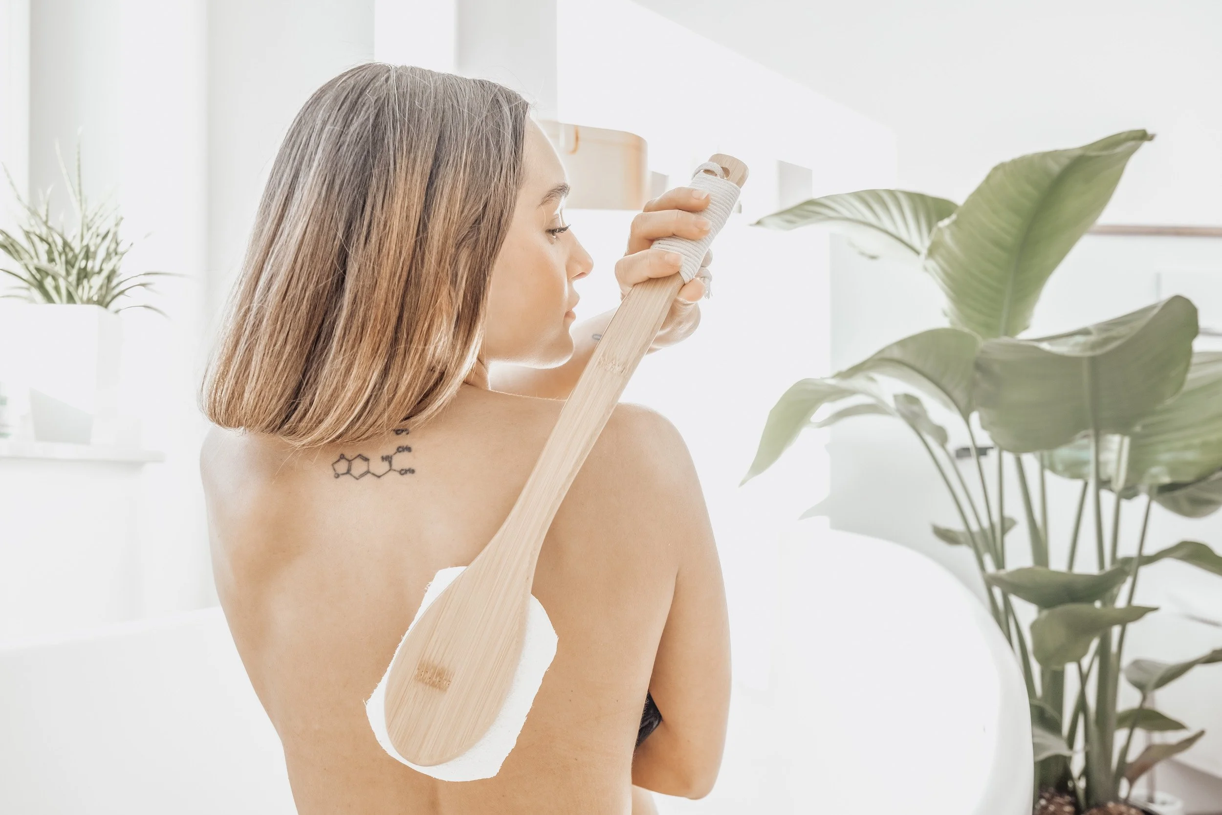 A woman with shoulder-length hair holding a large wooden spoon, using it as a back scrub brush, in a bright, minimalist bathroom with green plants.