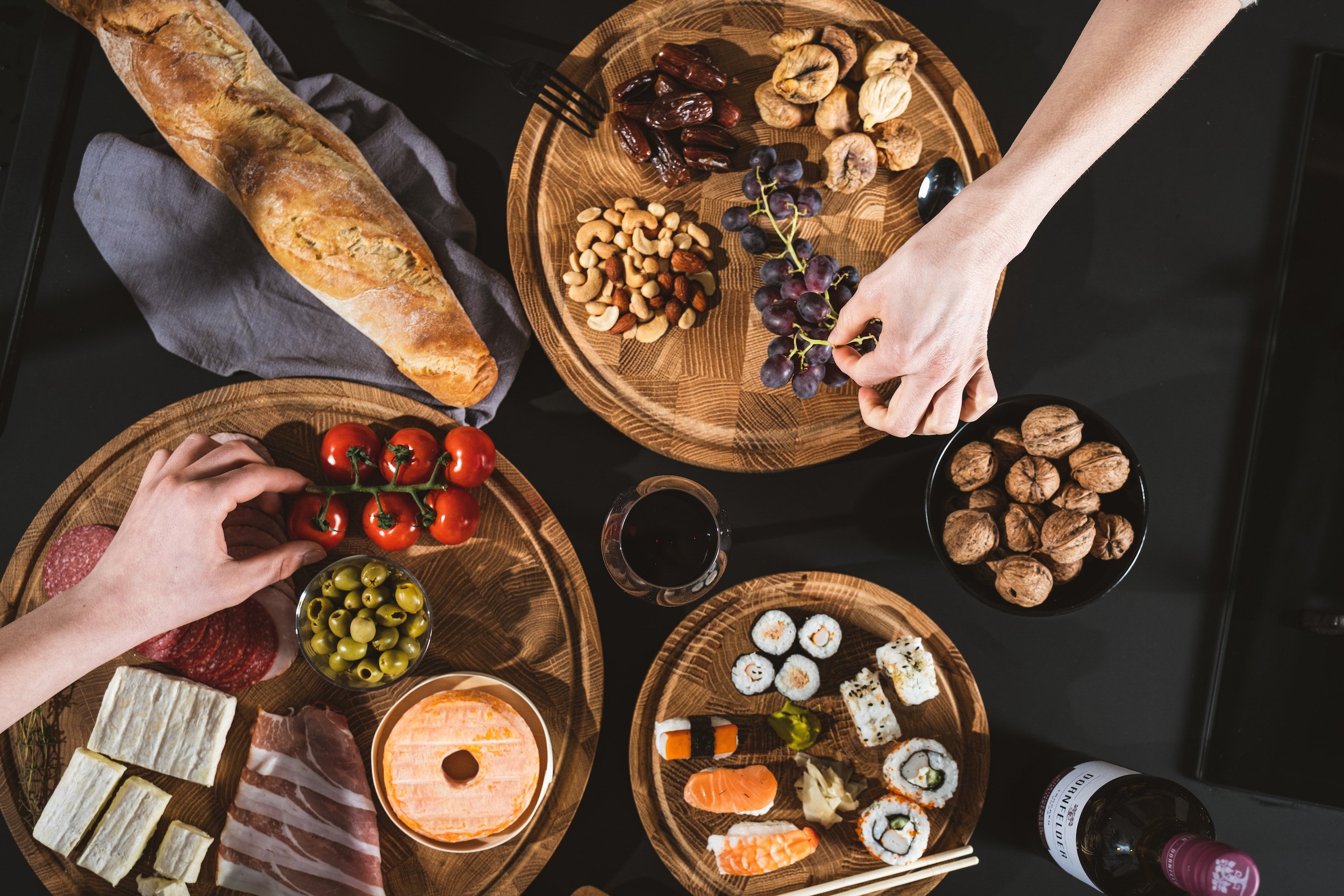 Overhead view of various snacks and foods on wooden plates, including a baguette, cherry tomatoes, green olives, sliced cheese, cured meats, various types of sushi, nuts, dried fruits, and a glass of red wine on a black table.