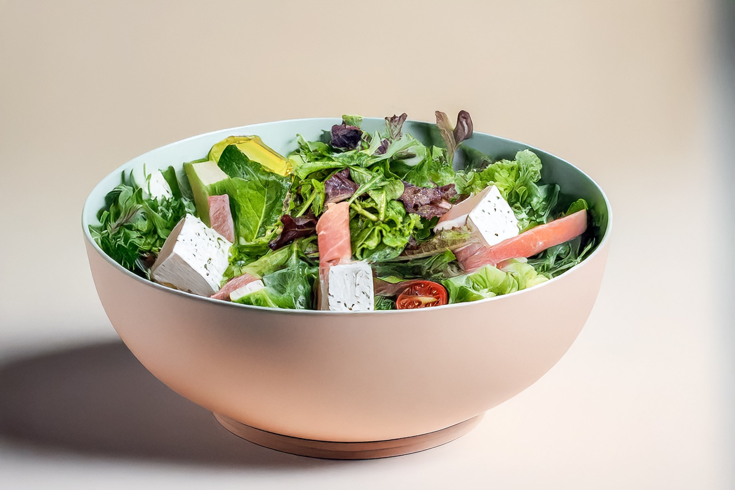 A bowl of mixed salad with greens, cherry tomatoes, cucumber, slices of ham, and cubes of white cheese.