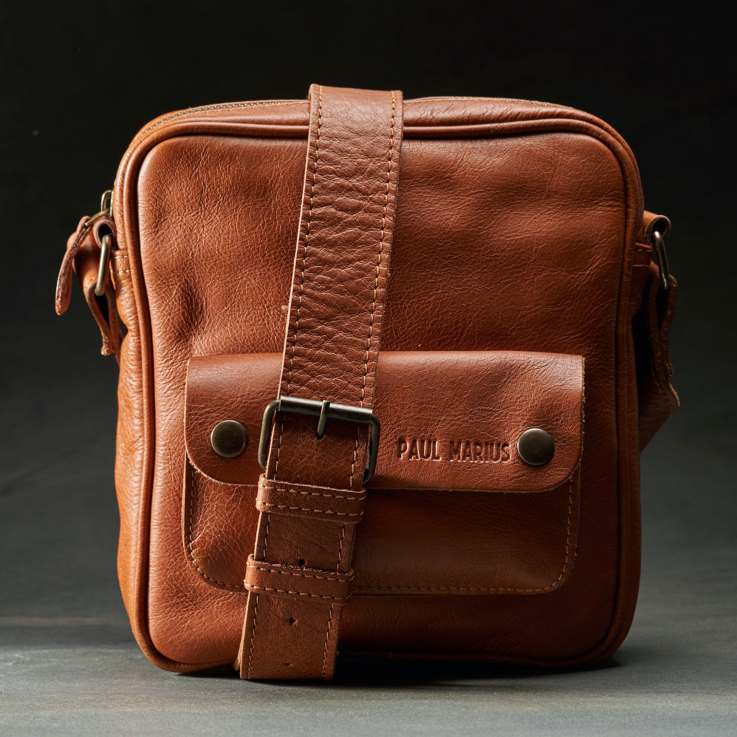 Brown leather shoulder bag with front pocket and adjustable strap, embossed with the name 'Paul Marius', against a dark background.