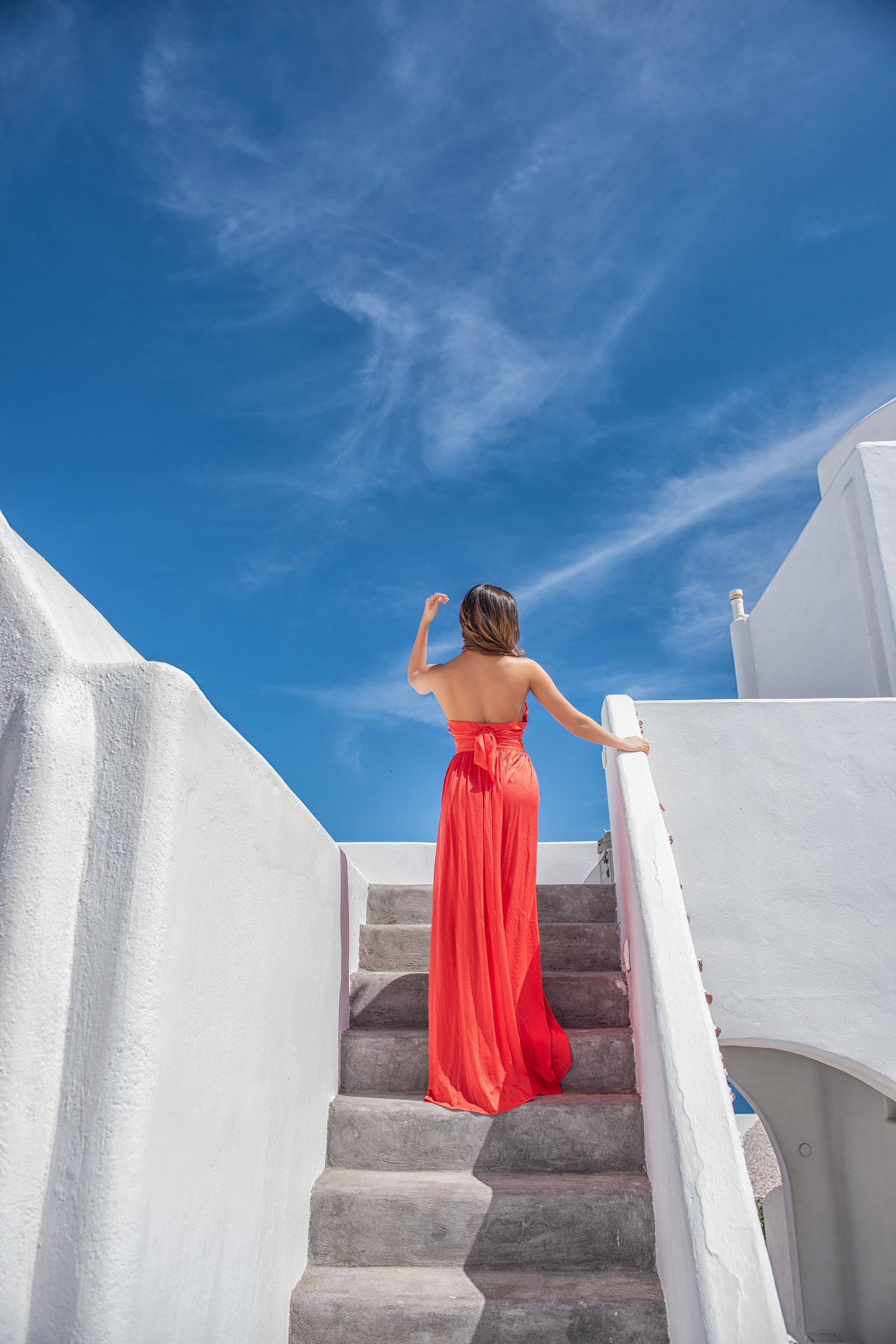 A woman in a strapless red dress standing on white stairs, facing away, against a bright blue sky with wispy clouds.