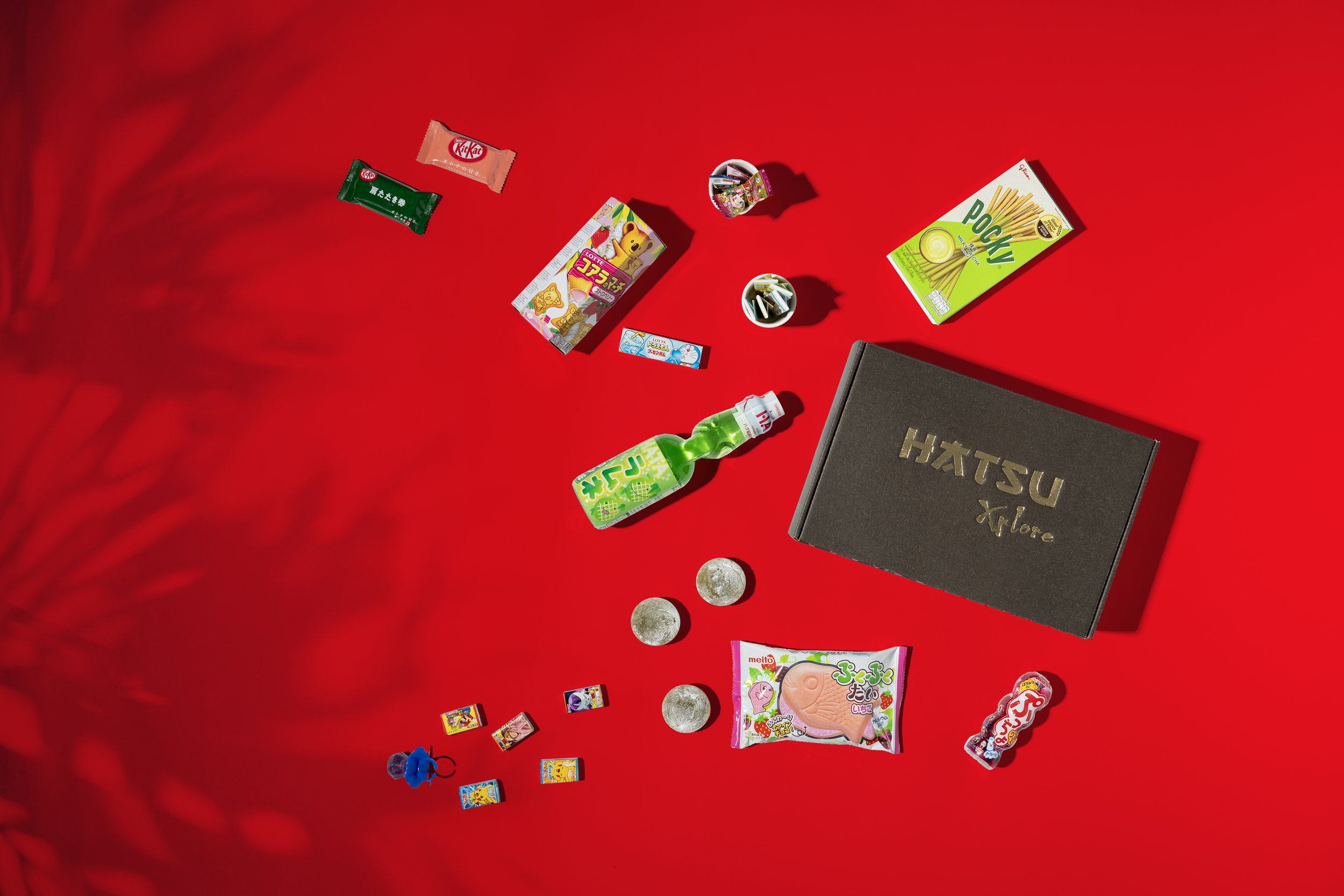 Assorted Japanese candies and chocolates scattered on a red surface, with a black box labeled "HATSU Xplore" among them.