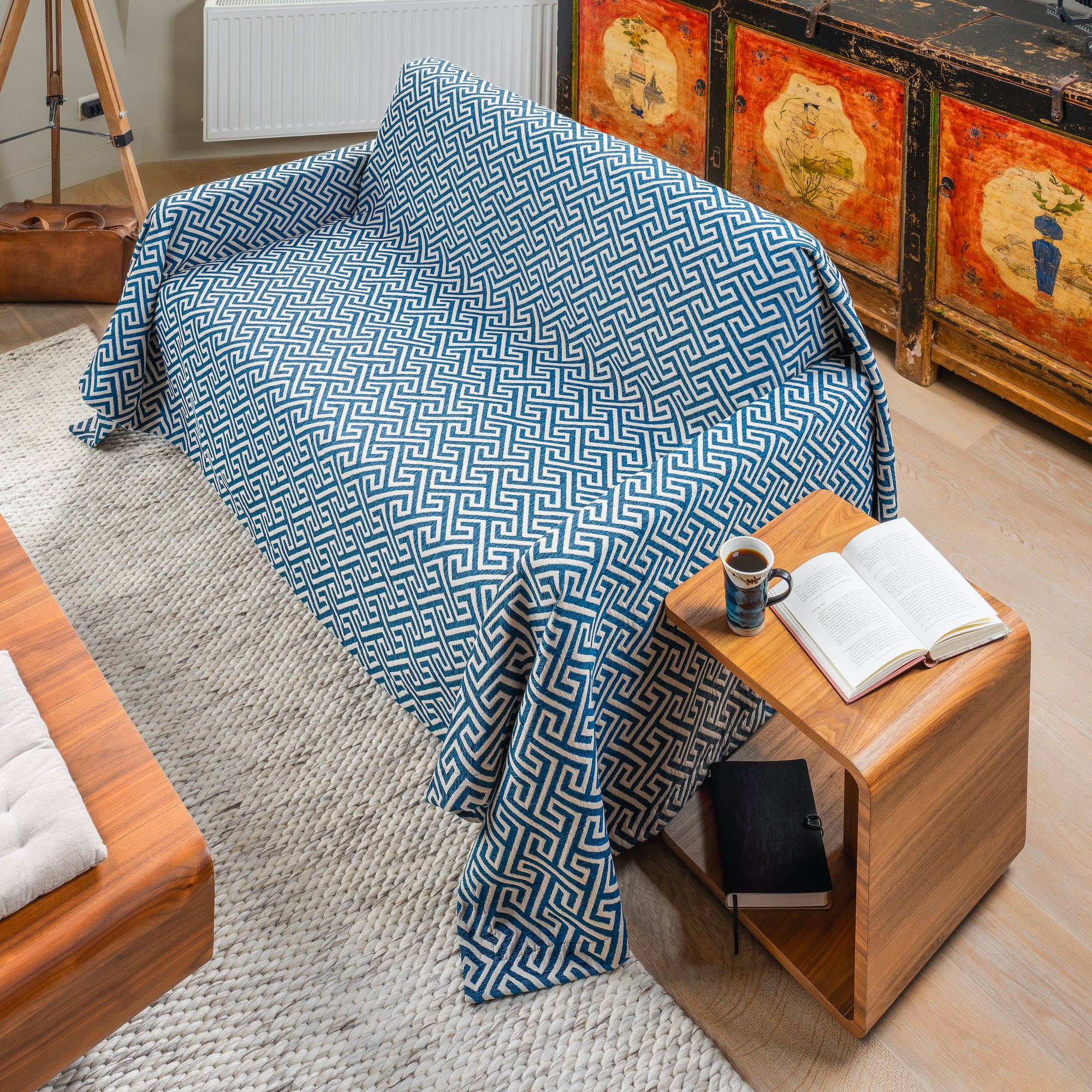 A cozy living room corner with a sofa covered in a blue and white geometric patterned throw. Beside the sofa is a small wooden side table holding an open book and a mug, with a black notebook underneath. In the background, a rustic cabinet with color
