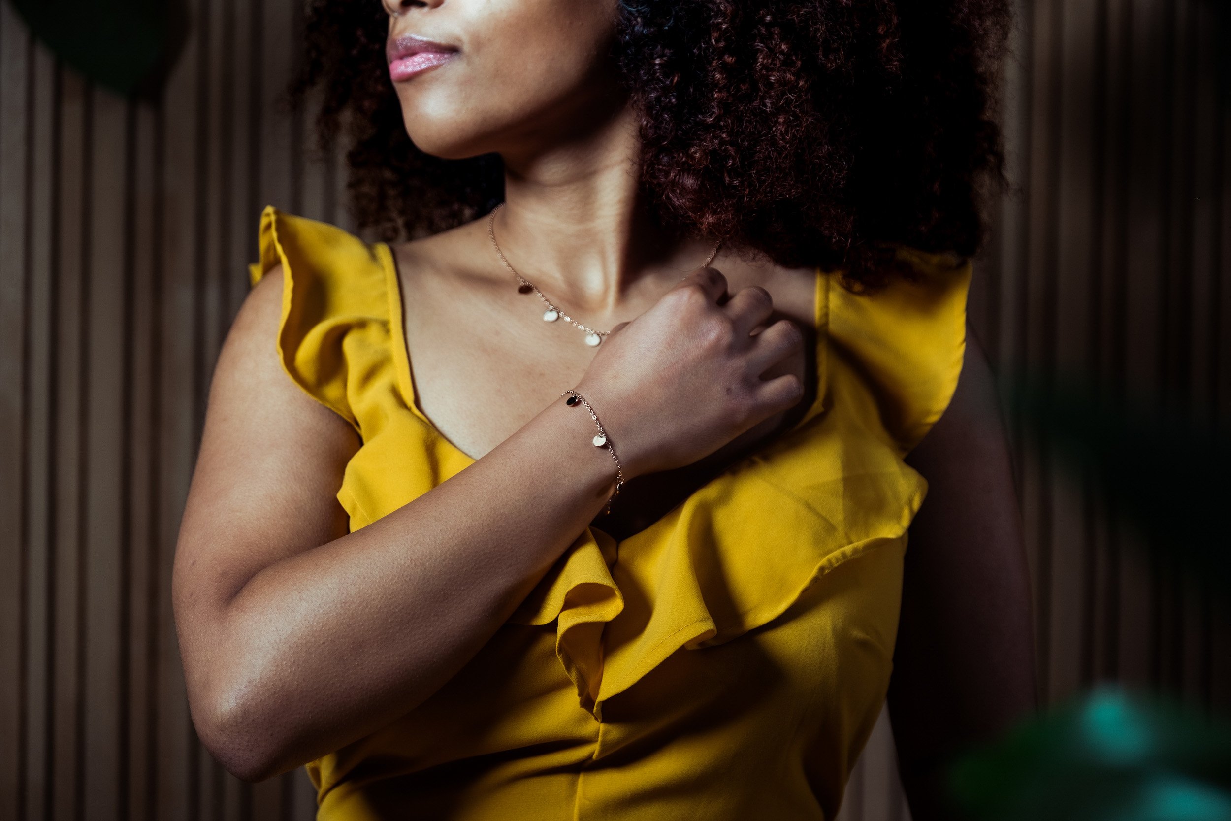 A woman with curly hair wearing a yellow dress, accessorized with jewelry, standing in front of a wooden background.