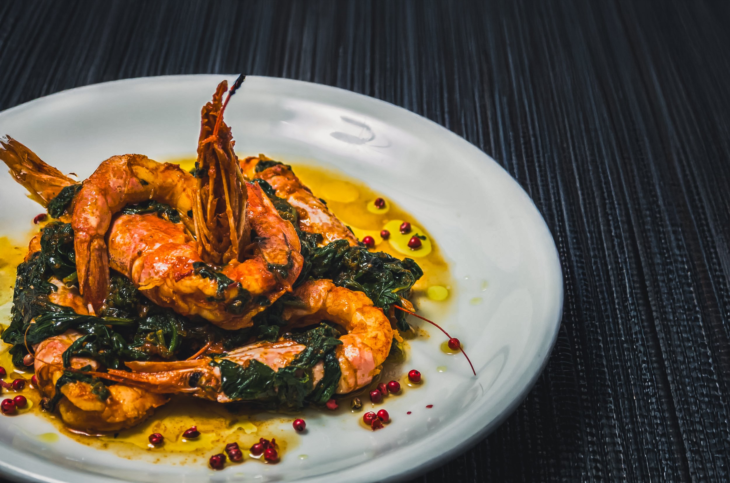 A plate of cooked shrimp with greens and a drizzle of sauce, garnished with red peppercorns and olive oil, on a white dish on a dark textured surface.
