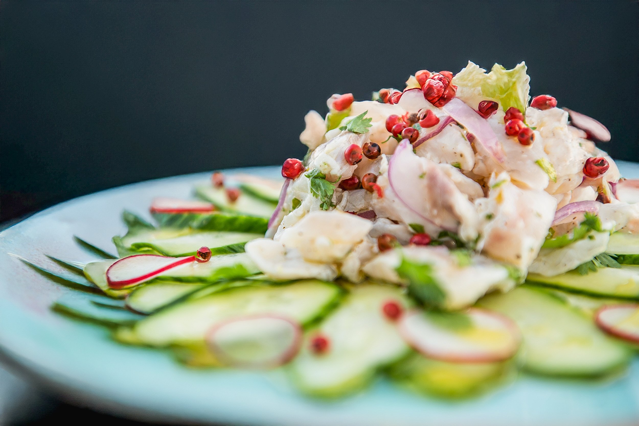 A plate of potato salad garnished with sliced radishes and red peppercorns, surrounded by thinly sliced cucumbers and herbs.