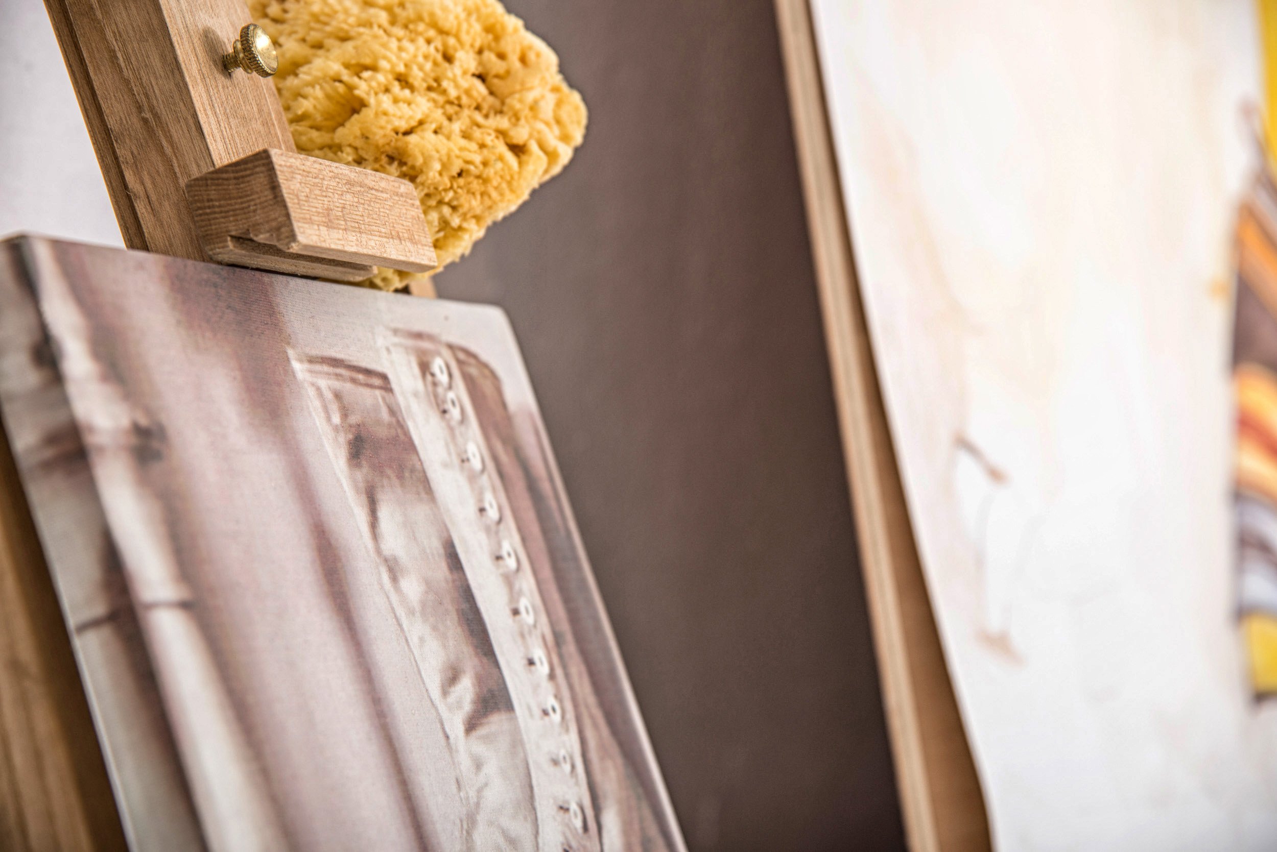 Close-up of a wooden art easel with a canvas, with a textured beige material on top of the easel, and other canvases in the background.