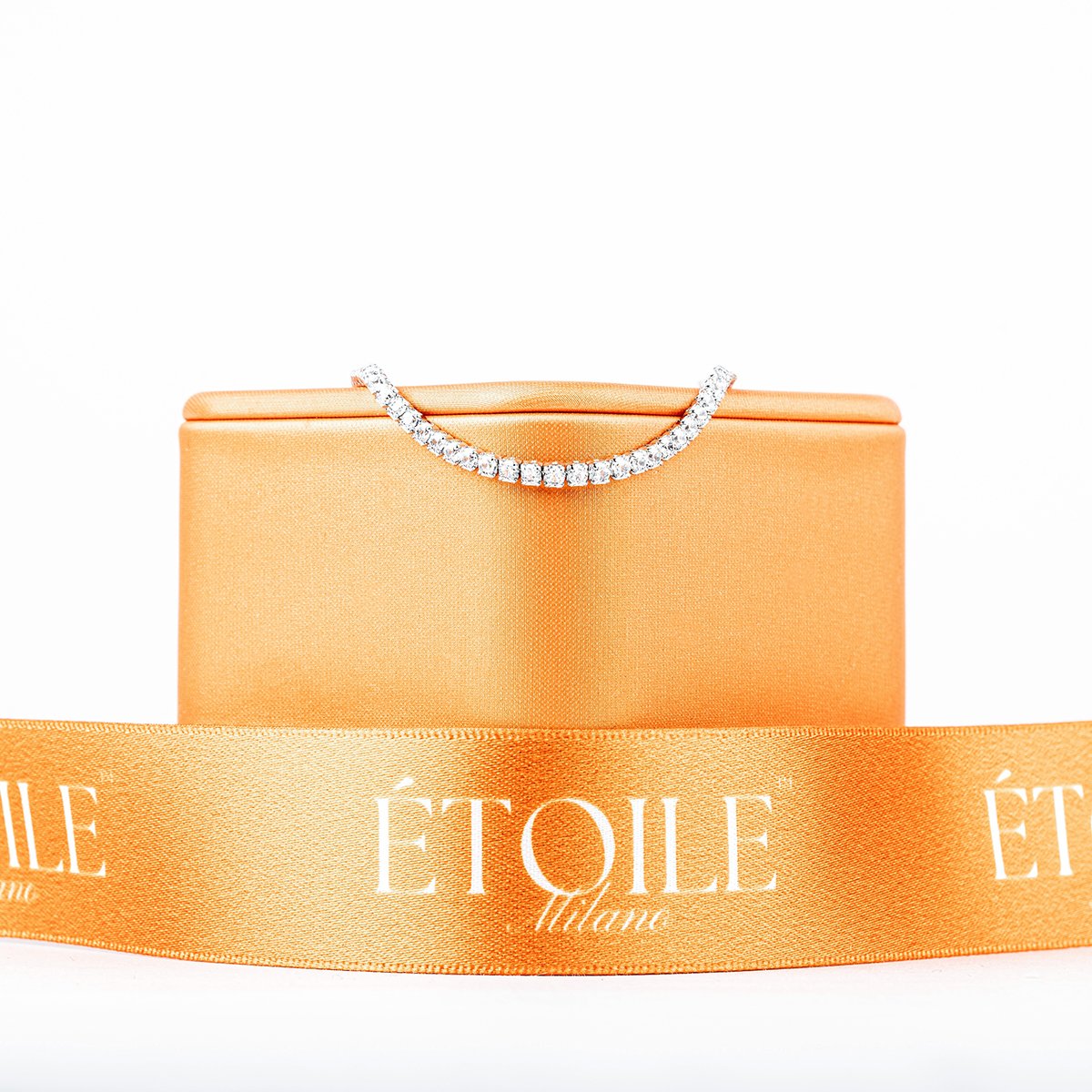 A diamond necklace draped over an orange jewelry box, with an orange satin ribbon in front reading 'Etoile' in white.