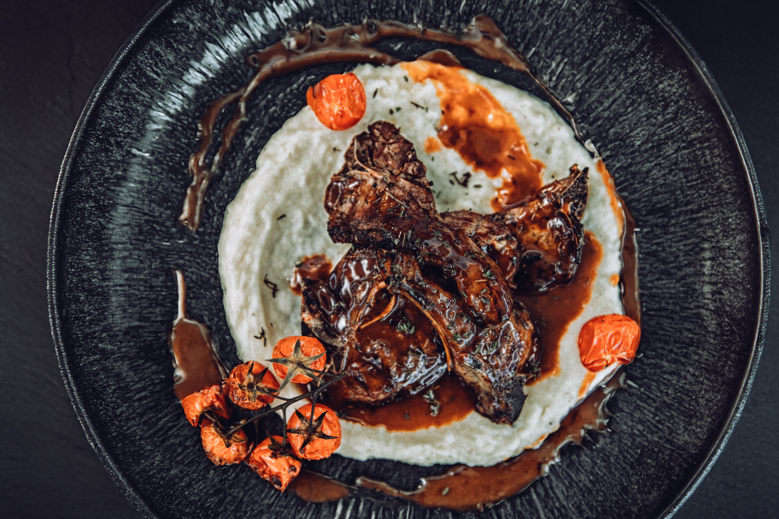 A black plate with a bed of mashed potatoes topped with braised ribs, garnished with cherry tomatoes and drizzled with dark gravy.