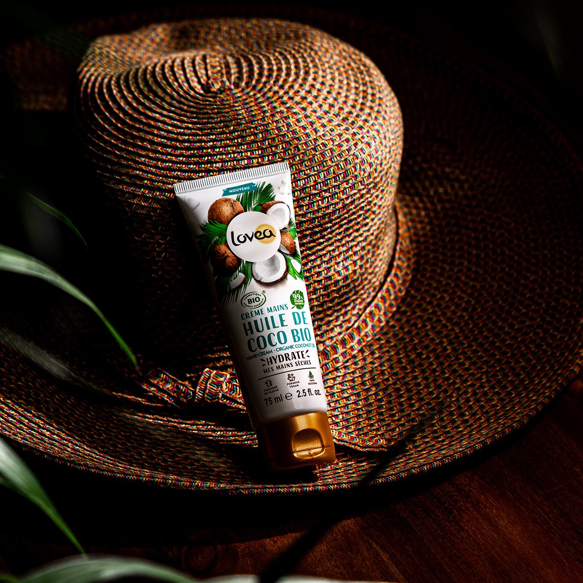 Sunlit colorful woven hat with a tube of Lovea organic coconut oil hand cream resting on it, surrounded by a dark background and a green leaf.
