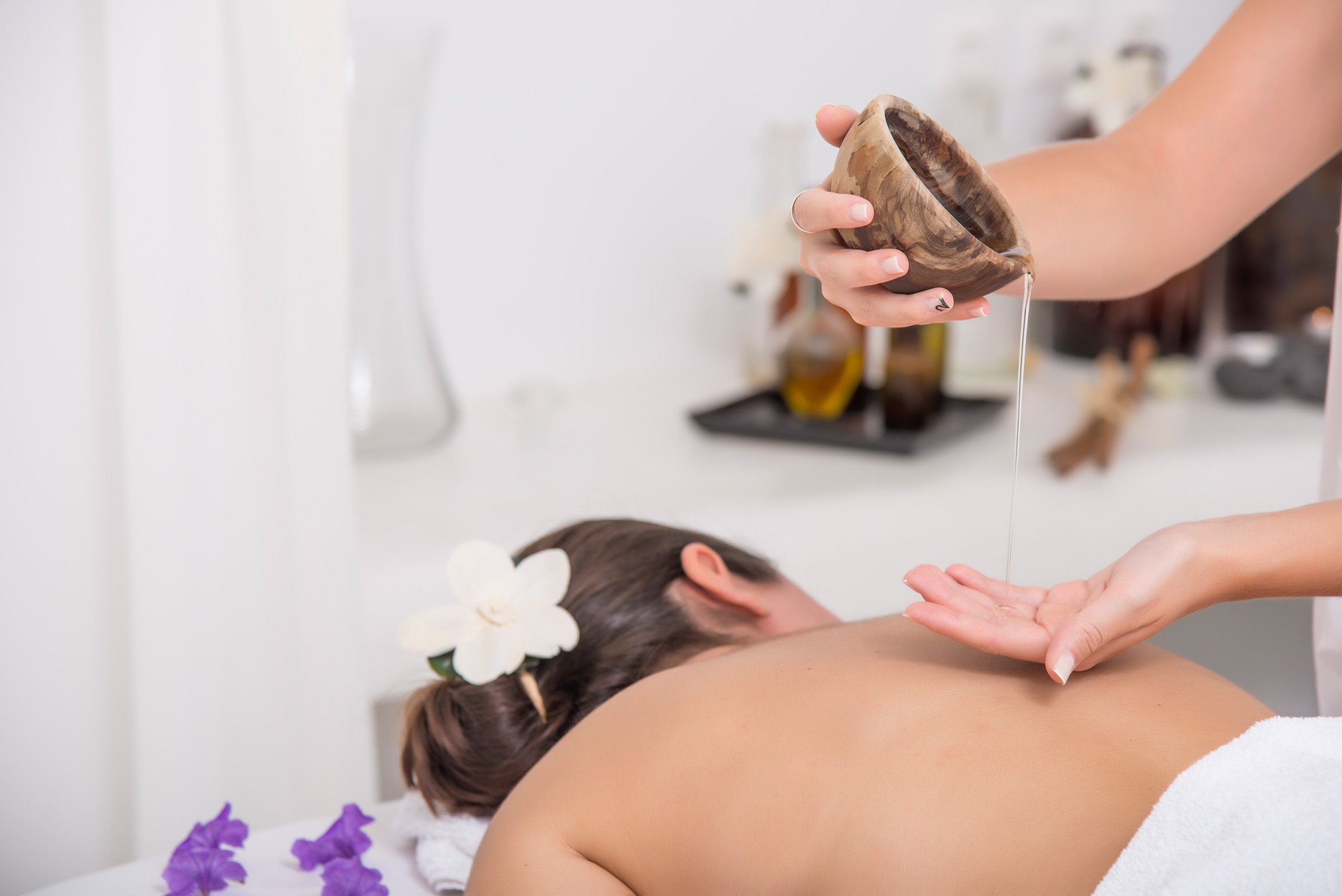 A woman receiving a massage with oil, lying face down on a massage table, with flowers in her hair, while a massage therapist pours oil onto her back from a wooden bowl.