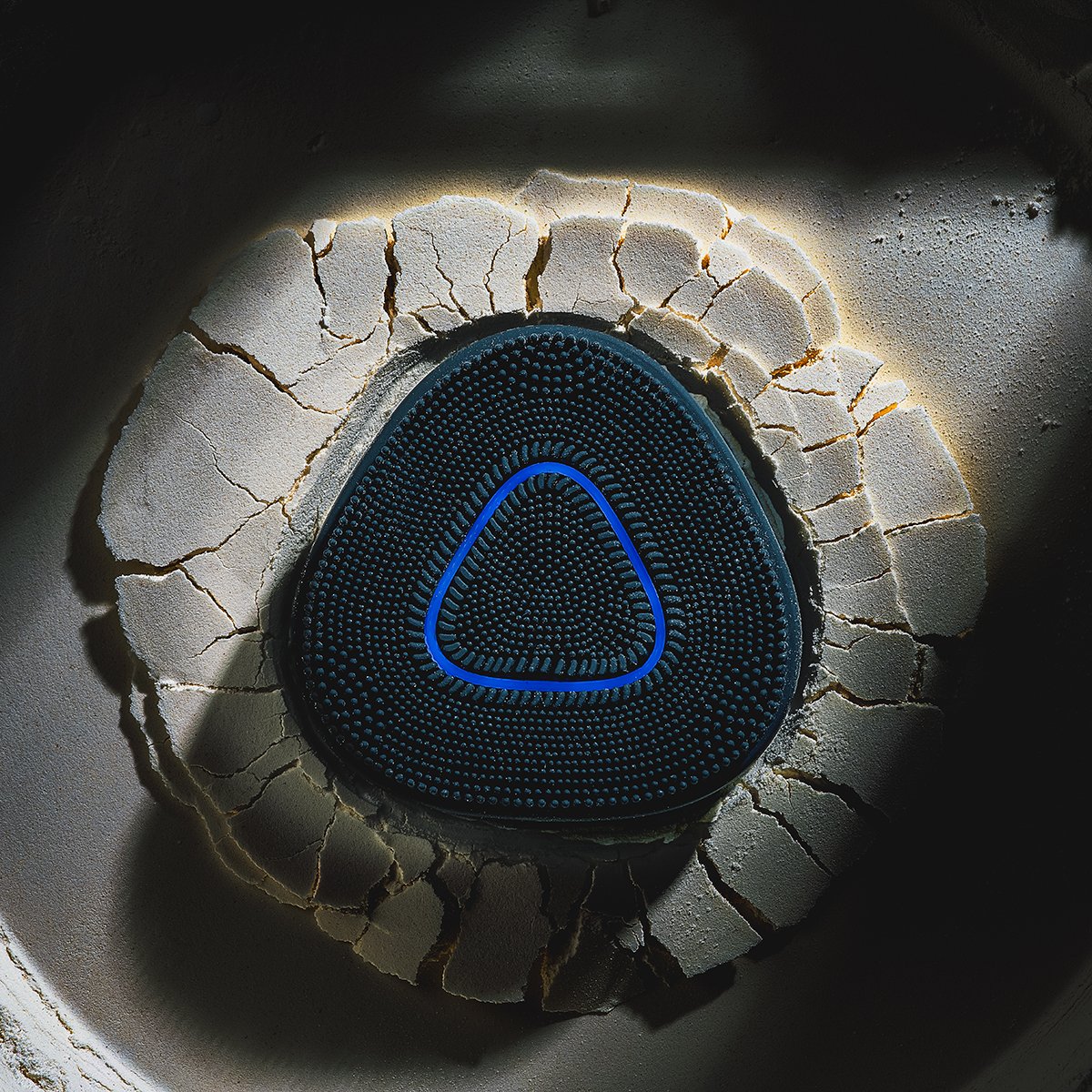Close-up of a black triangular wireless speaker with a blue outline, set inside a cracked, white ceramic bowl with lighting highlighting the cracks.