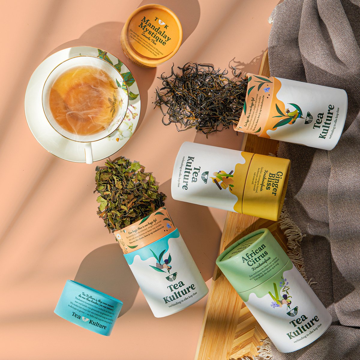 Assorted packages of Tea Kulture herbal teas, dried loose tea leaves, and a cup of brewed tea on a pink surface.