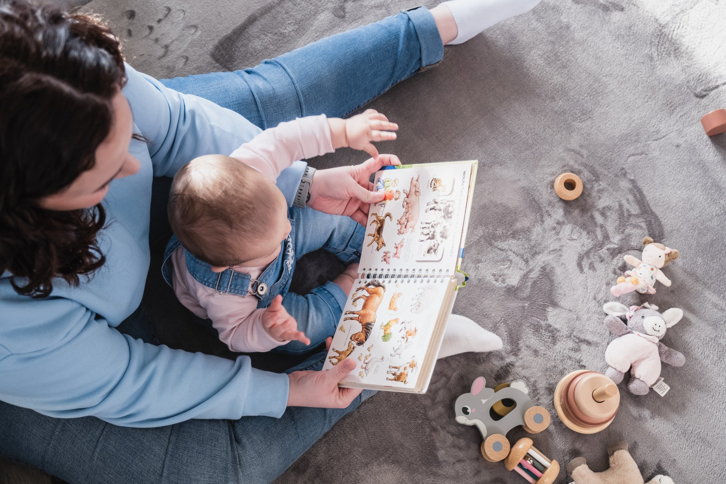 Adult and child sitting on gray carpet, reading a colorful animal book together. Around them are plush toys, wooden toys, and a rock tumbler.