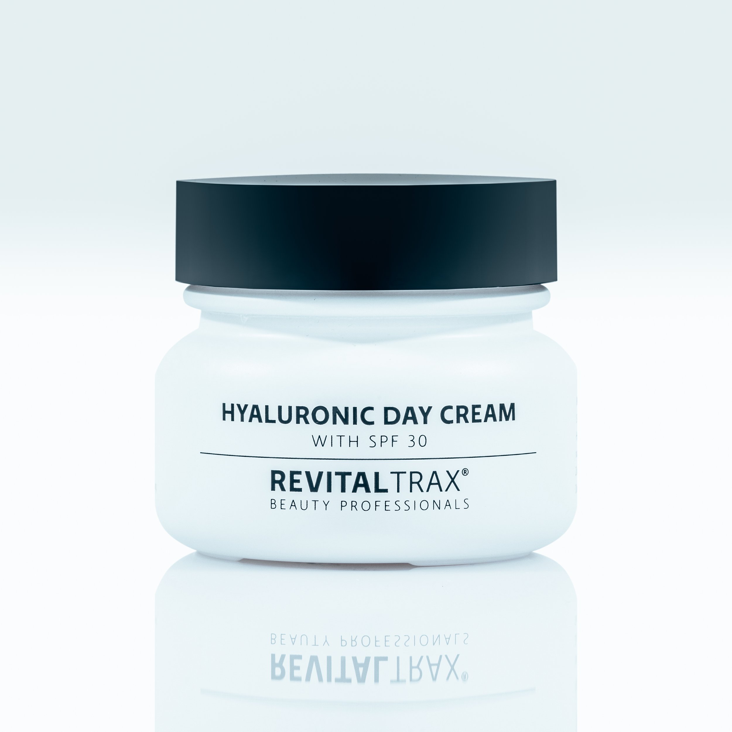 A white jar of Hyaluronic Day Cream with SPF 30 from RevitalTrax, with a black lid. The jar is reflected on a glossy surface.