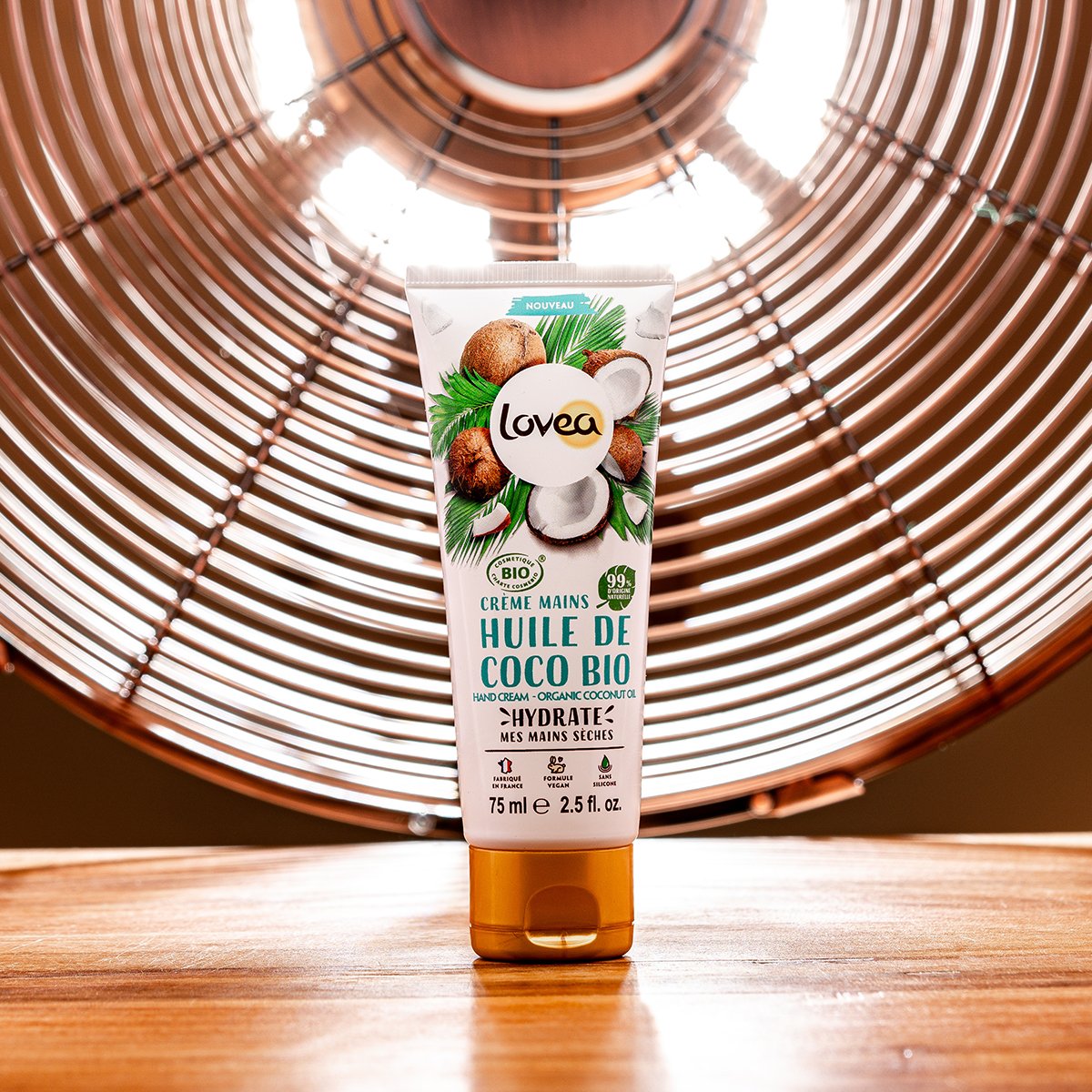 Tube of Lovea organic coconut oil hand cream placed on a wooden surface with a copper-colored fan in the background.