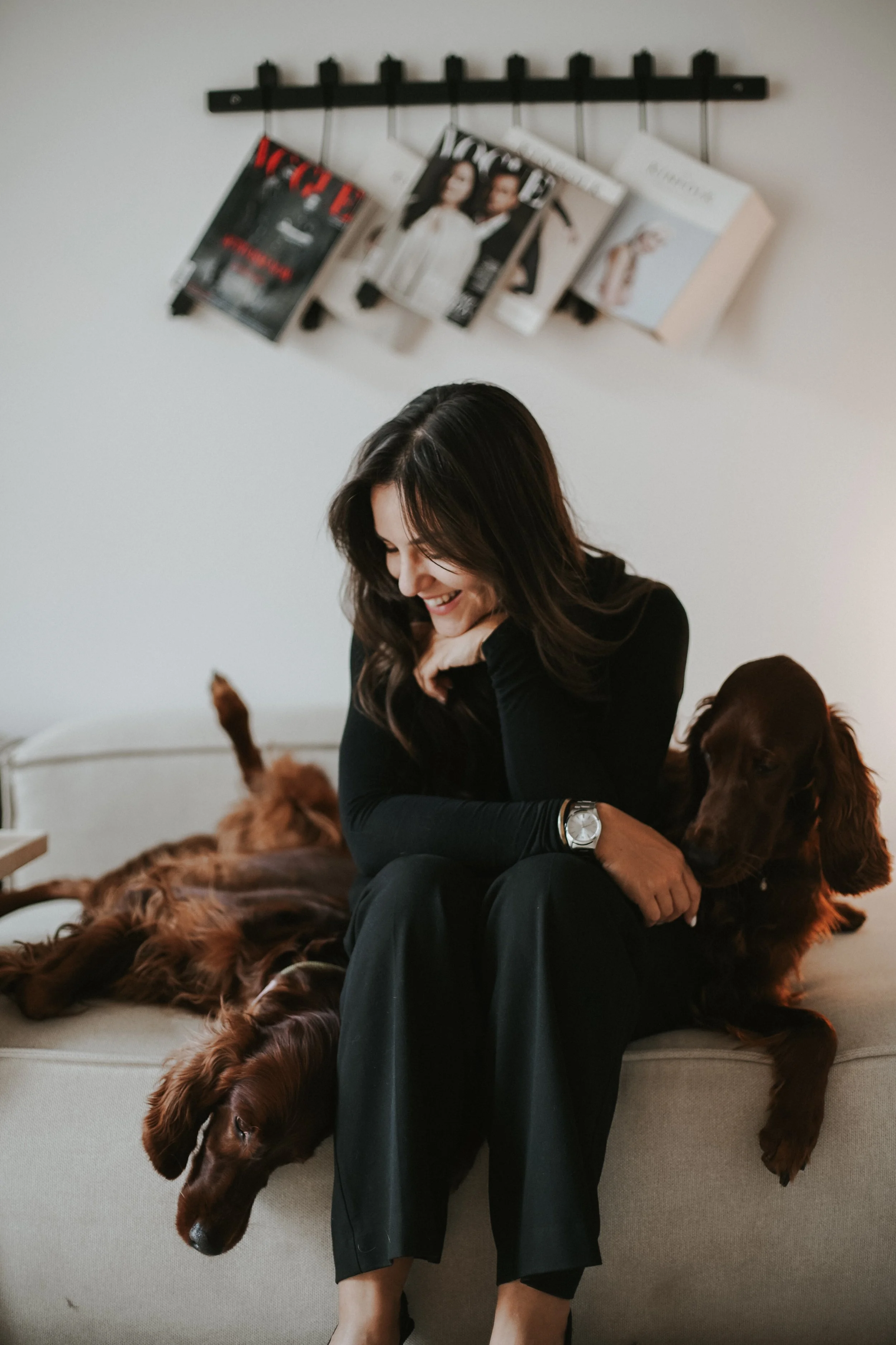 A woman sitting on a beige couch with two brown retriever dogs, one lying down with head hanging off the edge and the other sitting close beside her, smiling and looking down as she interacts with the dogs. There are magazines hanging on a black rack