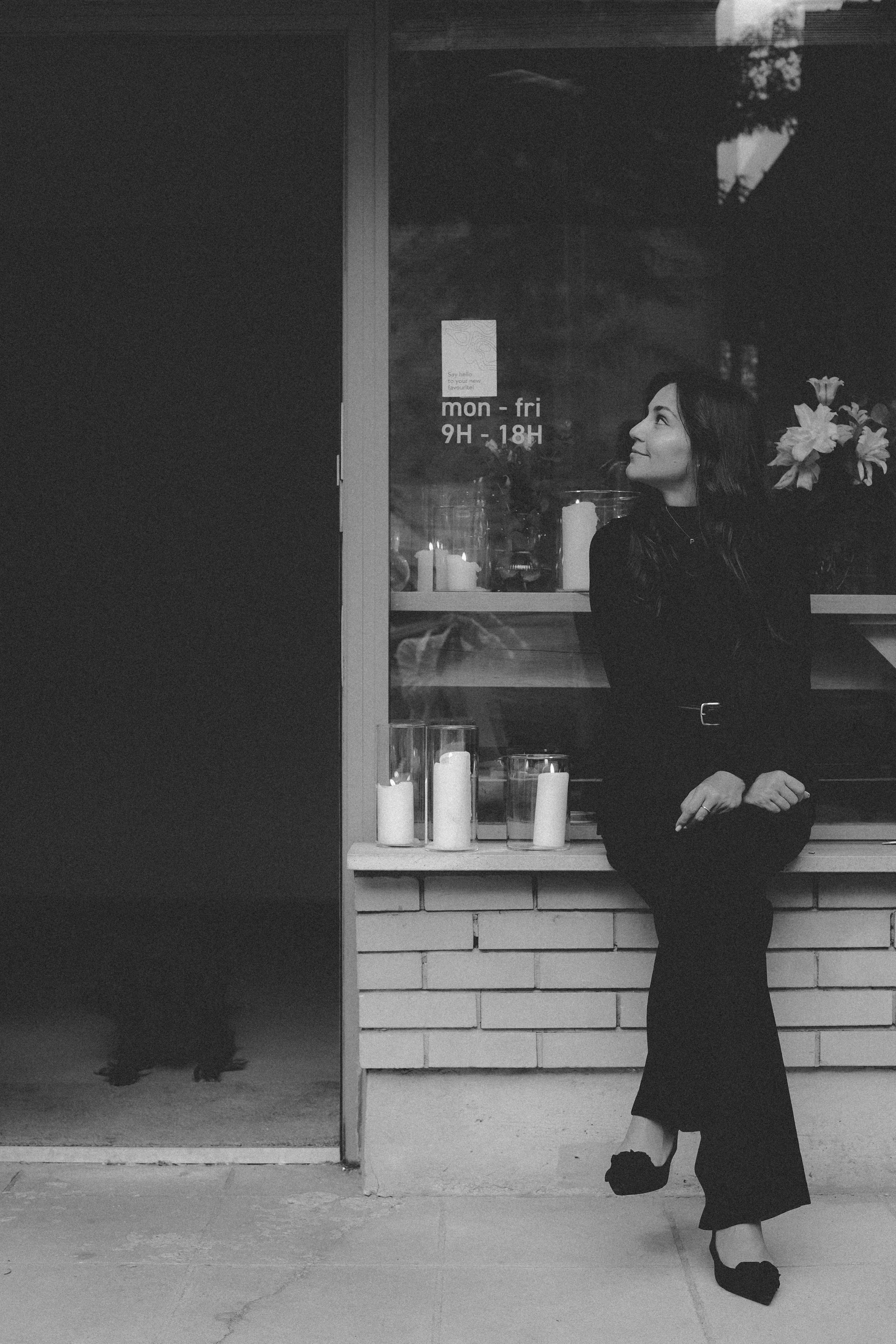 A woman sitting on a window sill outside a shop, looking to the right. She is dressed in black, with candles in glass holders placed beside her. The shop has a sign showing business hours from Monday to Friday, 9 AM to 6 PM.