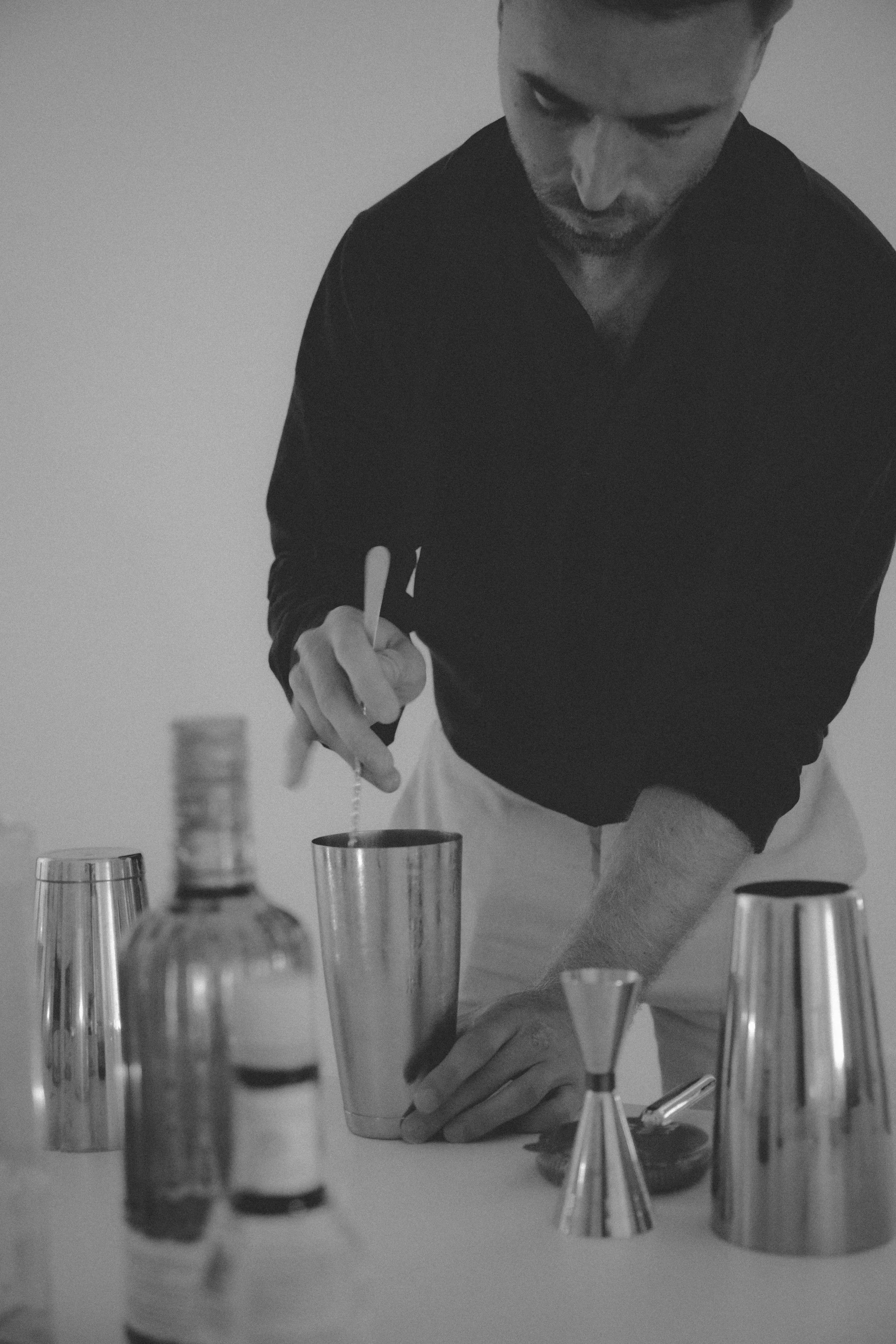 Man preparing a cocktail, pouring liquid into a shaker in a bar or kitchen setting, with various bottles and jiggers on the table.