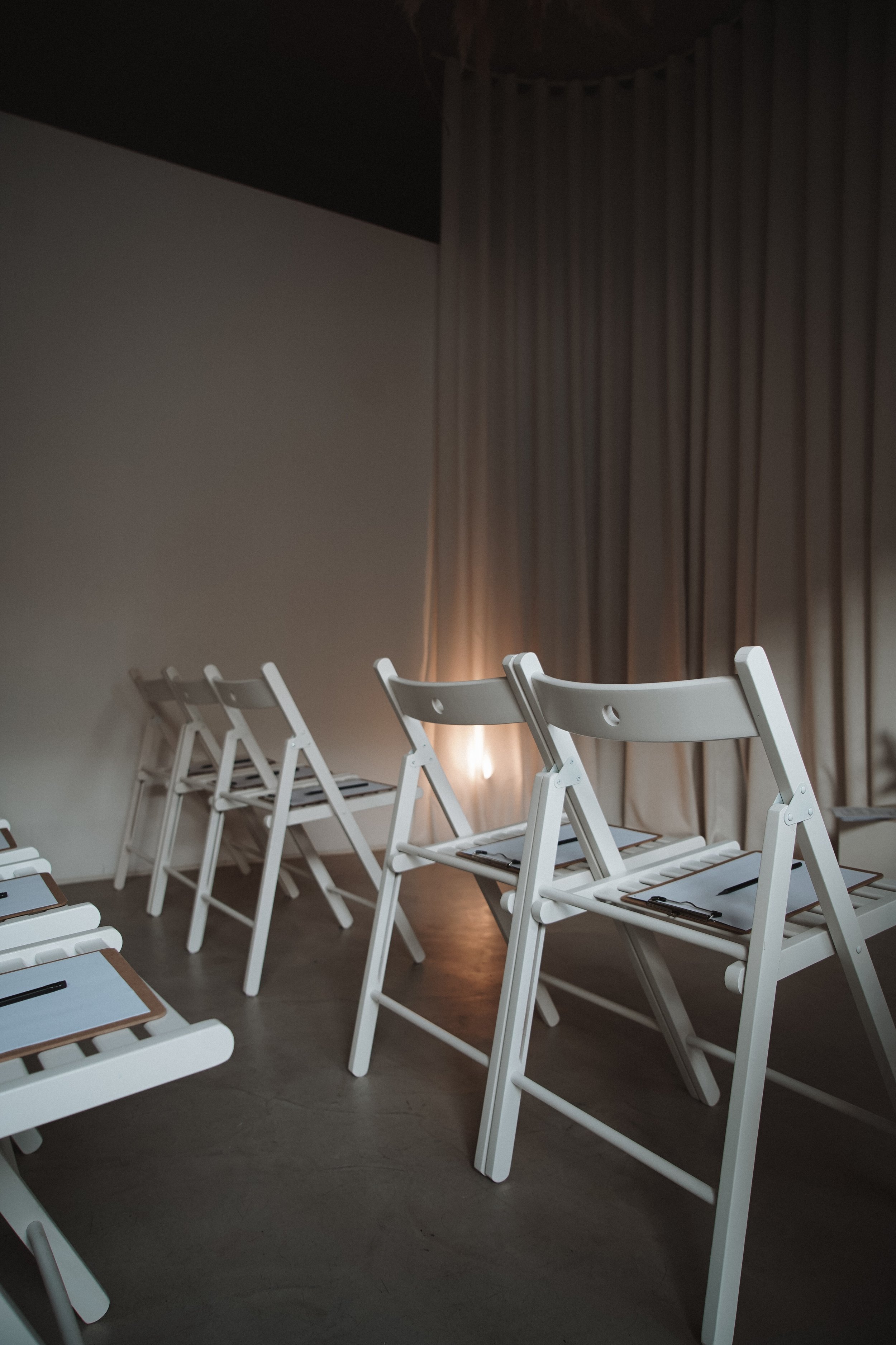 Empty white folding chairs with clipboards and pens arranged in rows in a dimly lit room with curtains.