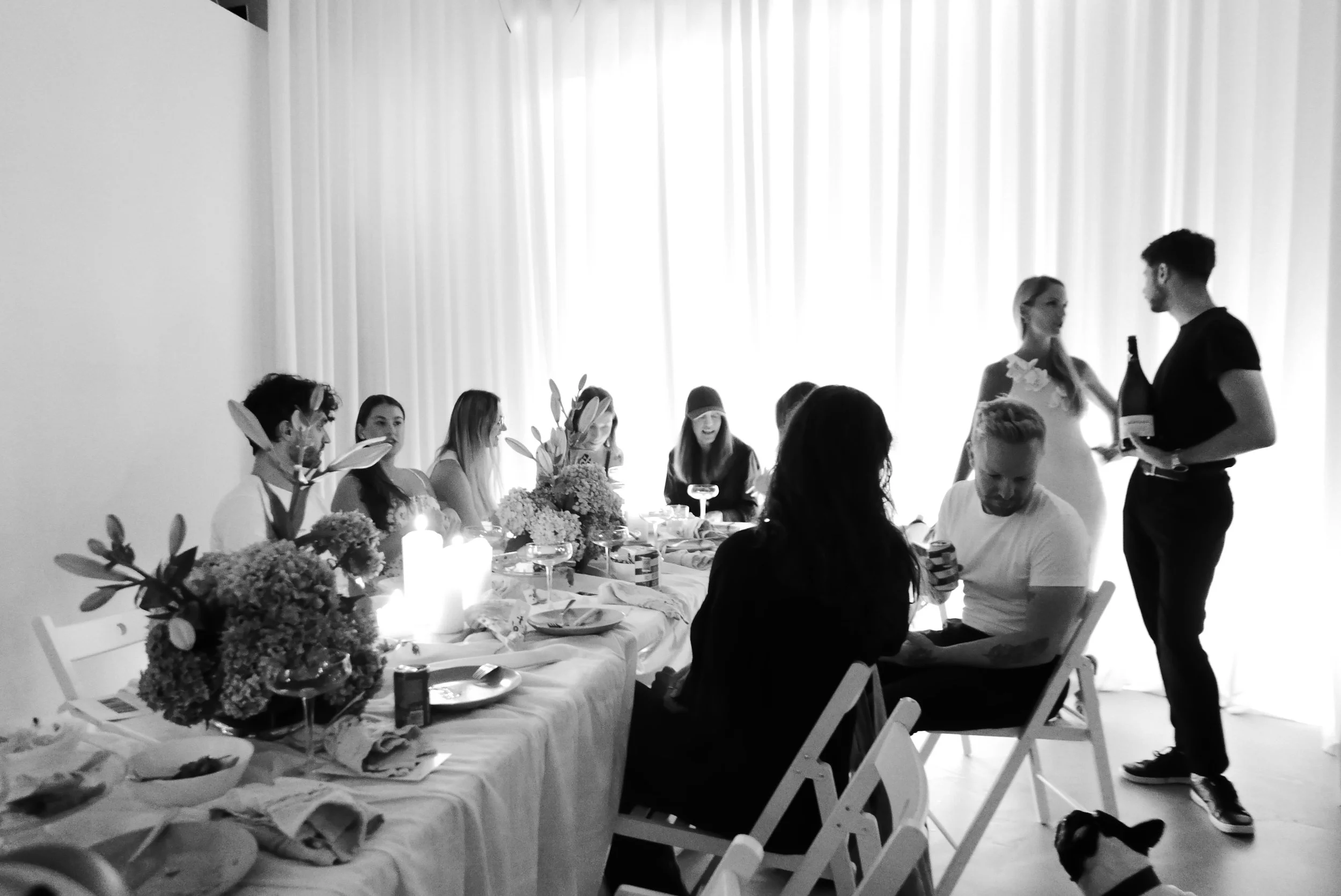People gathered at a table during a celebration, with some talking and others sitting, decorated with flowers and candles, with two individuals standing and exchanging a bottle.