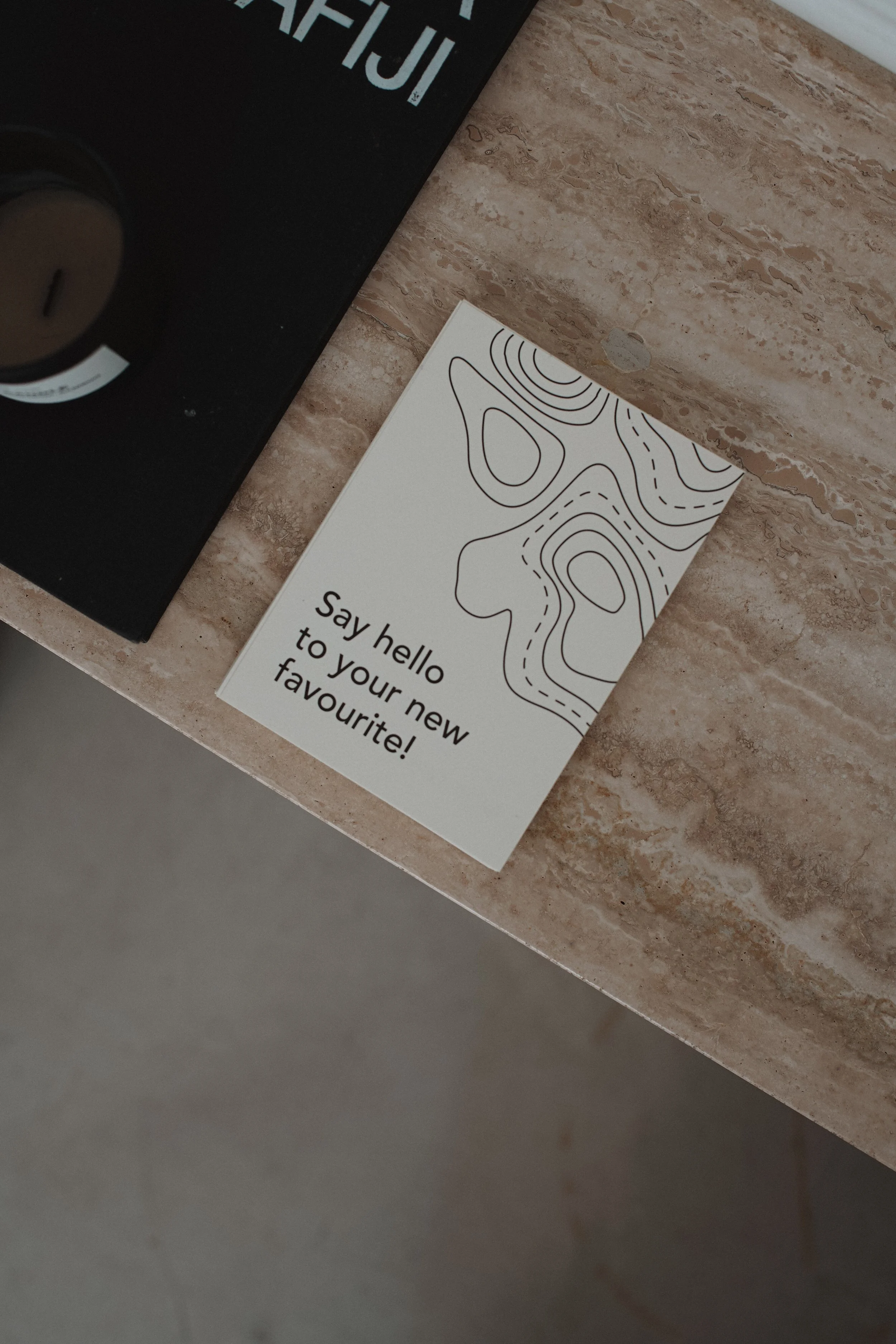 A white card with abstract contour lines and the text "Say hello to your new favourite!" placed on a beige marble countertop next to a black box with partially visible white text and a candle.