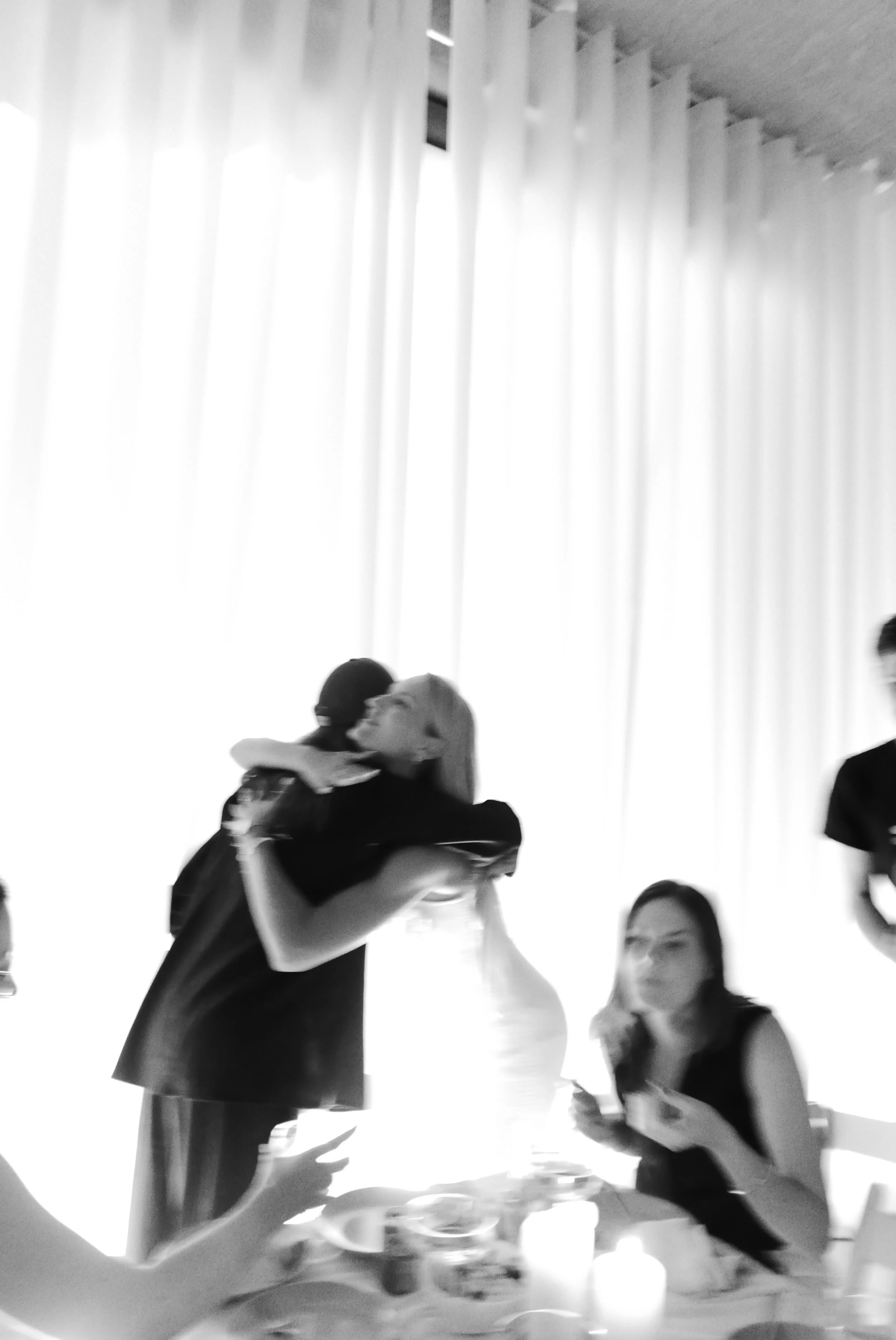 Two women hugging at a celebration or gathering, with a table of food and drinks in front of them, and curtains in the background.