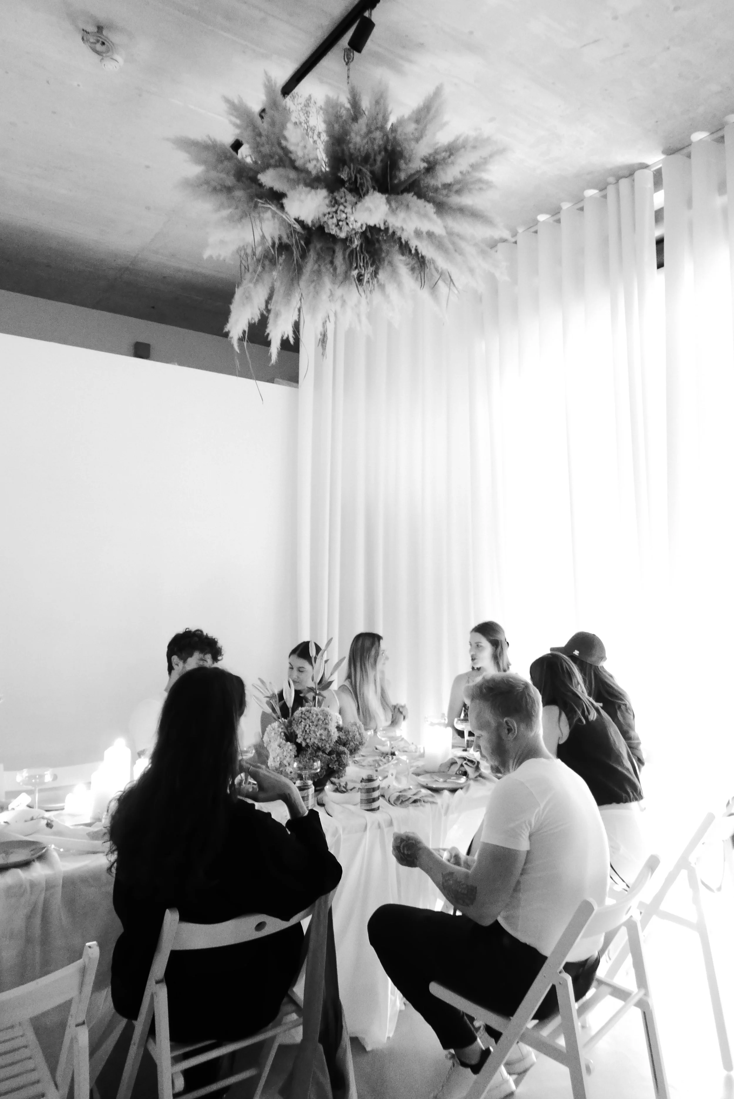 People gathered around a dining table with floral centerpieces, engaging in conversation during a meal or celebration, with a large pampas grass arrangement hanging from the ceiling.