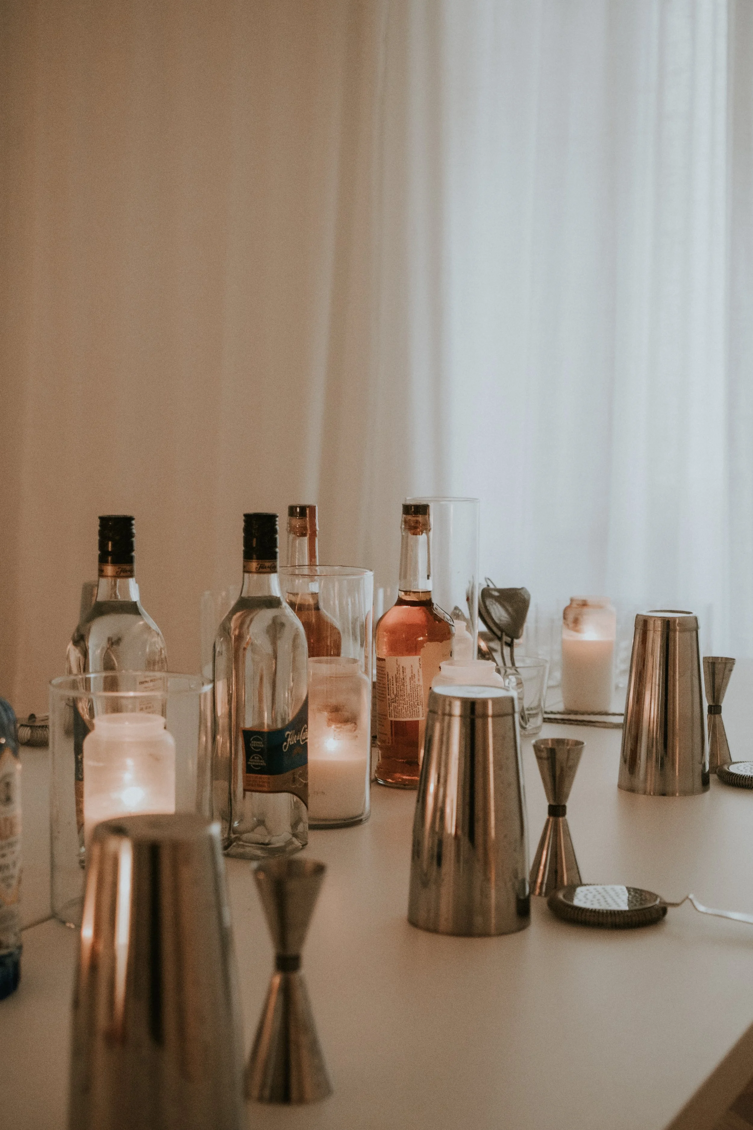 Table with bottles, candles, cocktail shakers, and bar tools in a softly lit room.