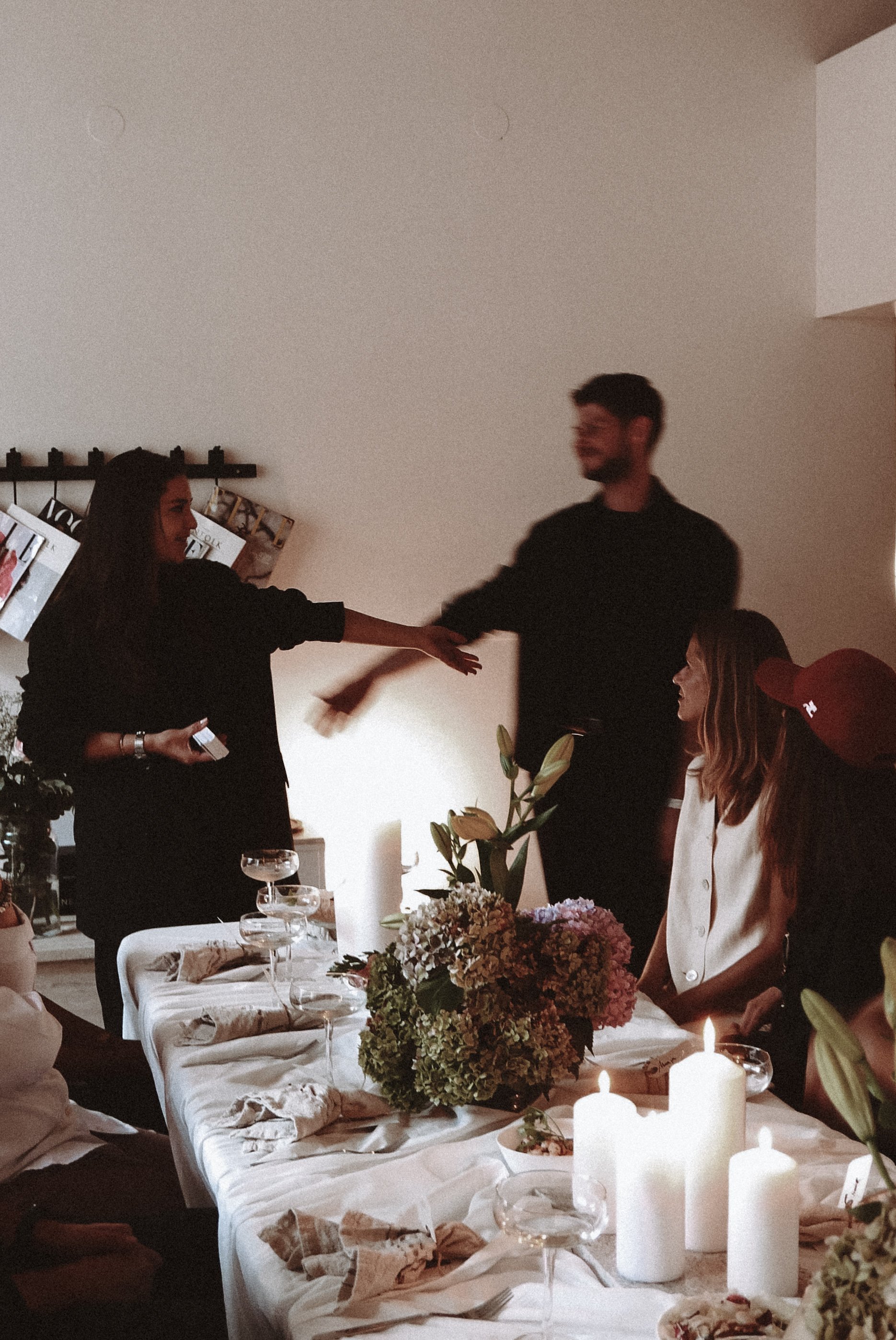 People gathered around a dining table with floral centerpieces and candles, with two people dancing and others sitting and watching.