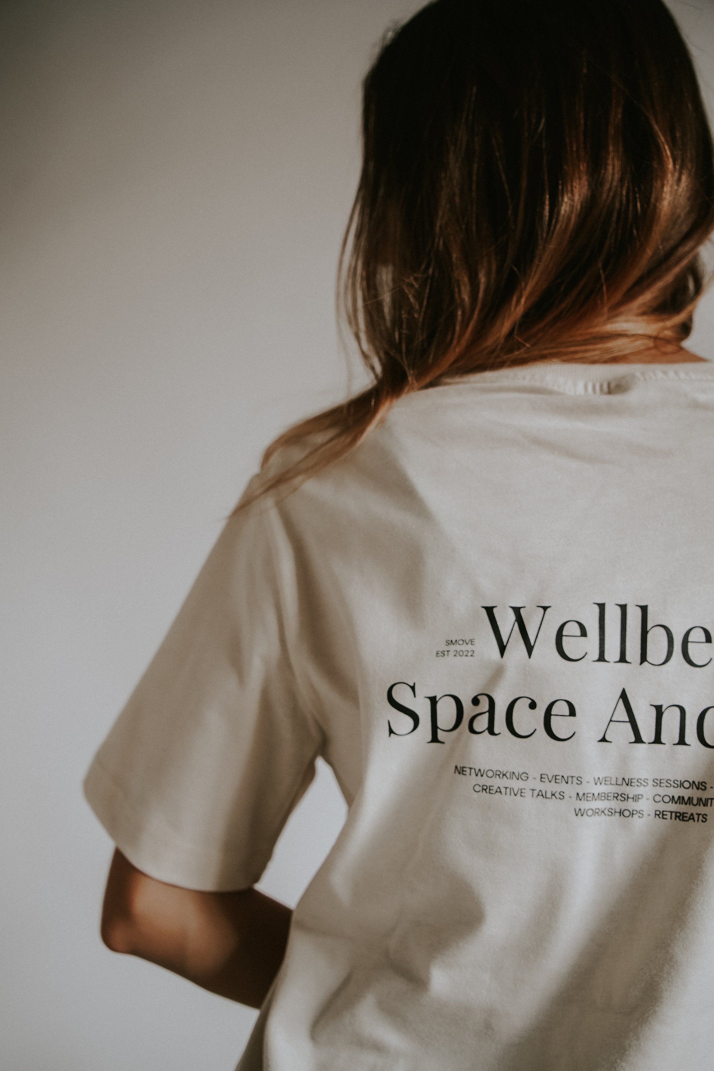 Person with brown hair wearing a beige t-shirt with black text about 'Wellbe Space And' and event details.